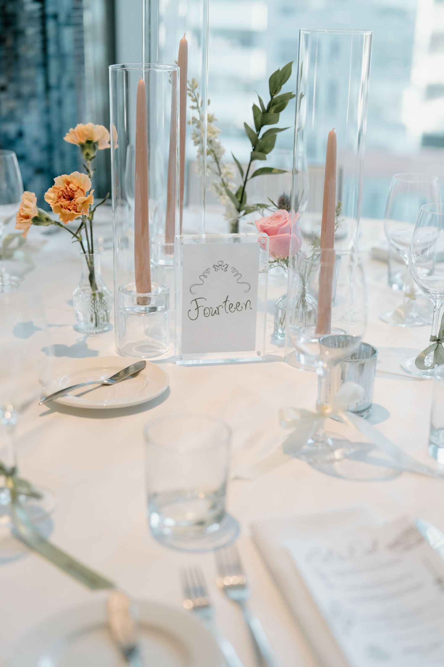 Elegant wedding table setting with tall glass vases, pink and peach flowers, and table number displayed on white linens.
