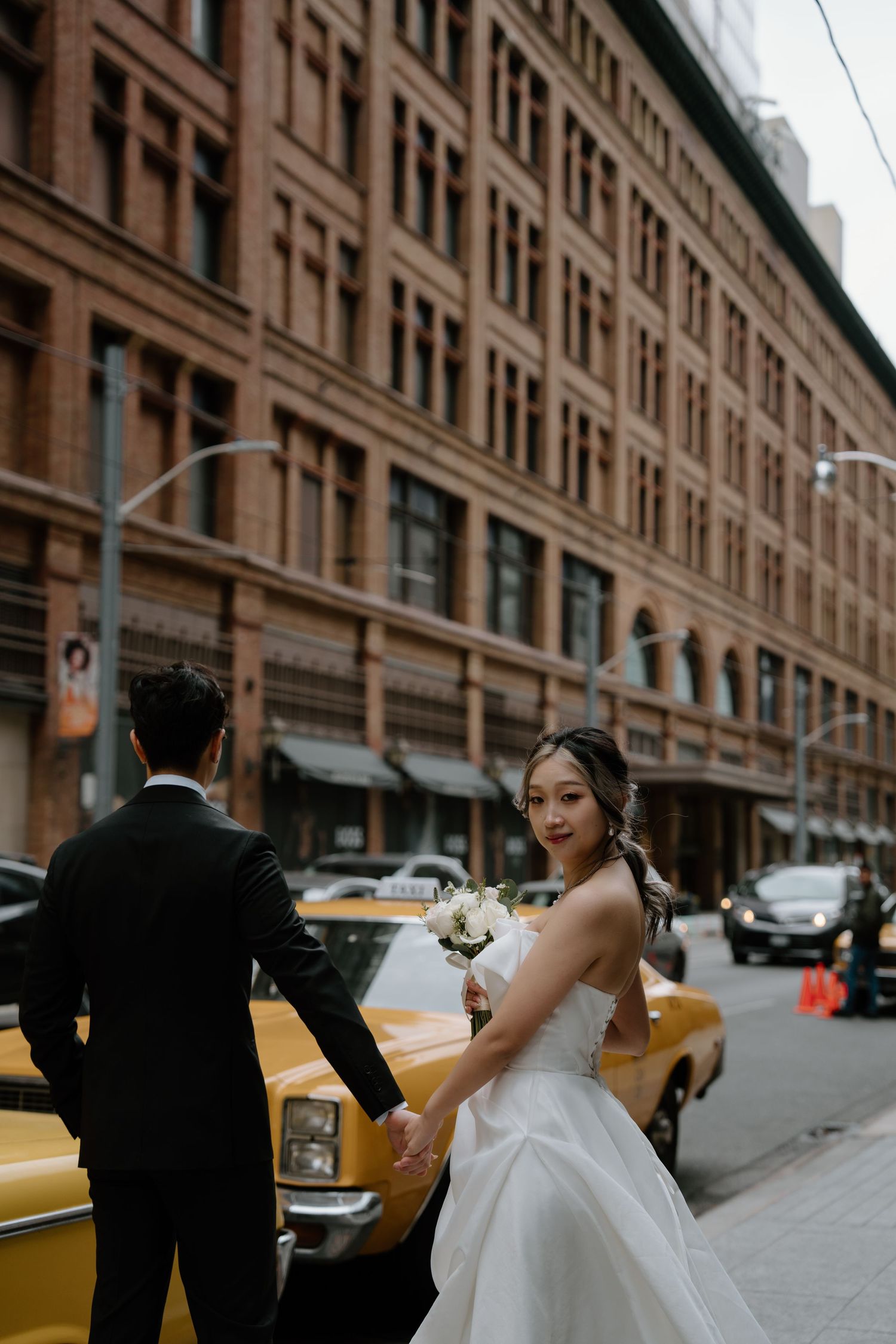 A bride and groom pose next to a yellow taxi cab on a city street with historic buildings.