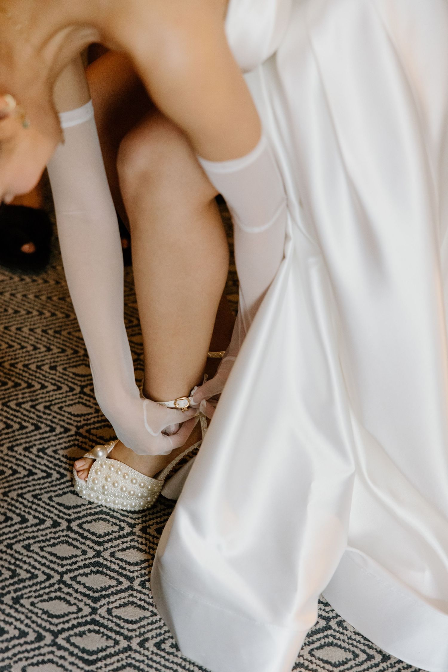 White bridal shoes and stockings being put on against a geometric patterned carpet during wedding preparations.