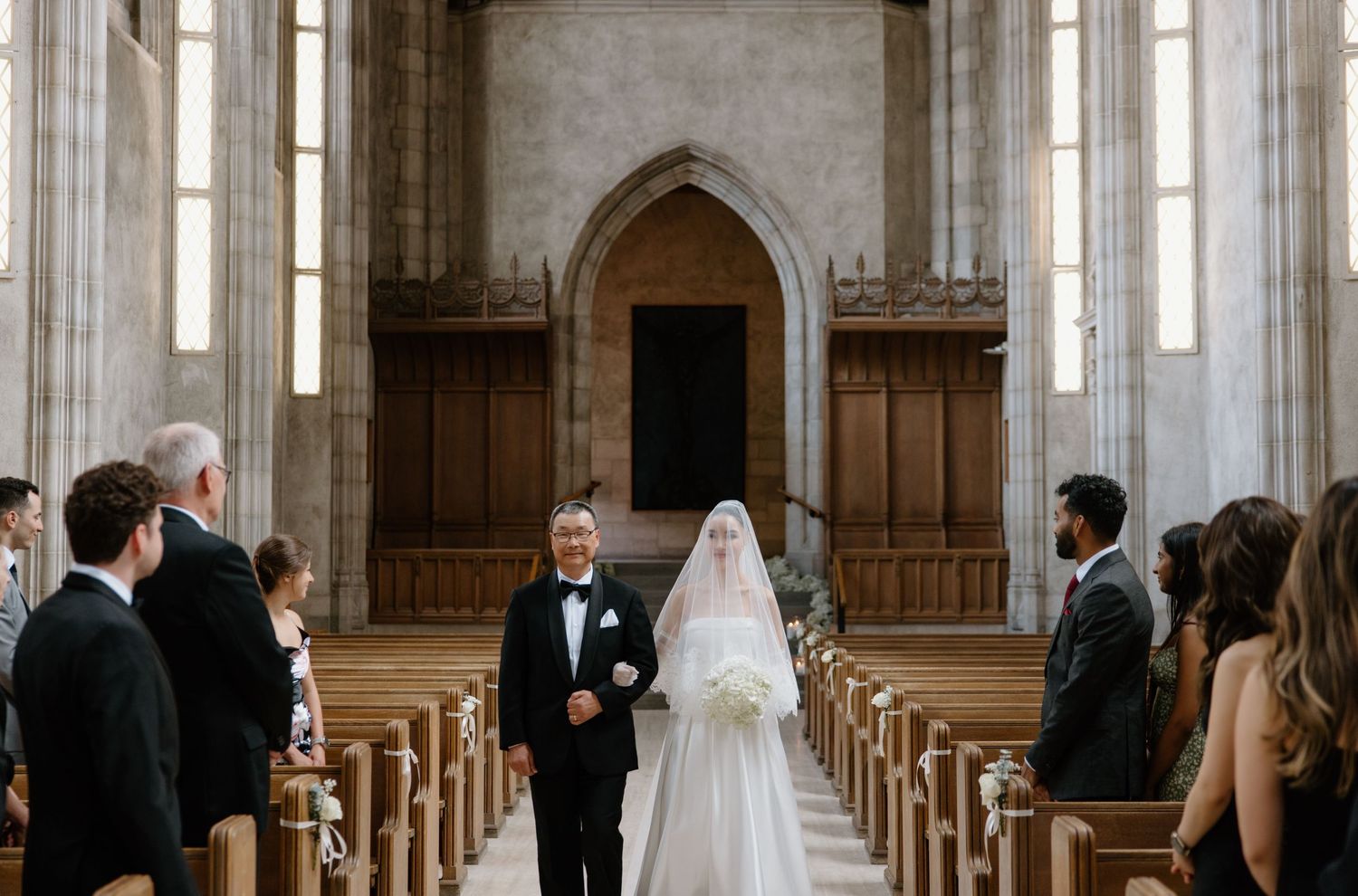 Inside a gothic church with arched windows and wooden pews during a formal wedding ceremony.