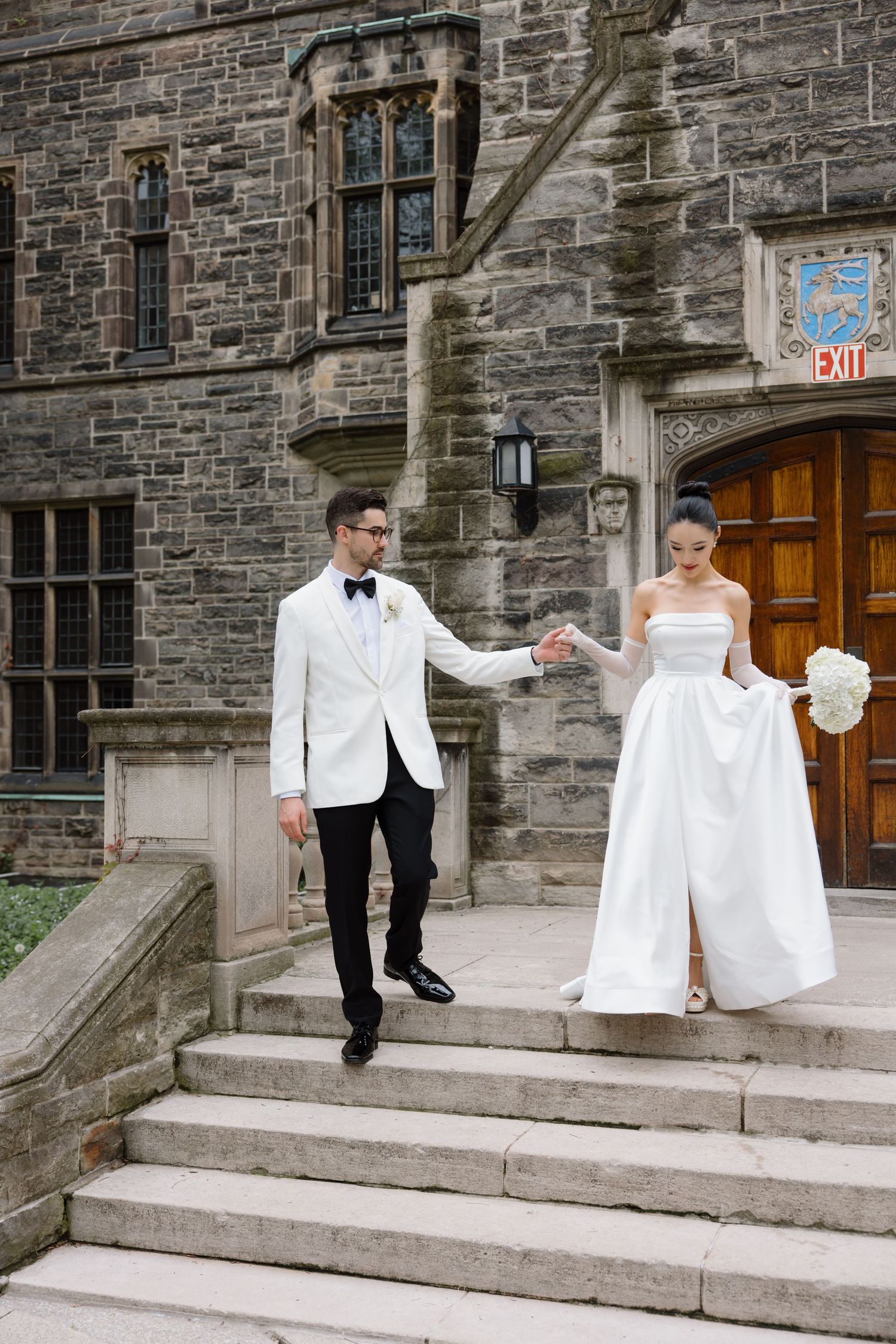 A couple in wedding attire descends stone steps outside a gothic style building with stained glass windows.