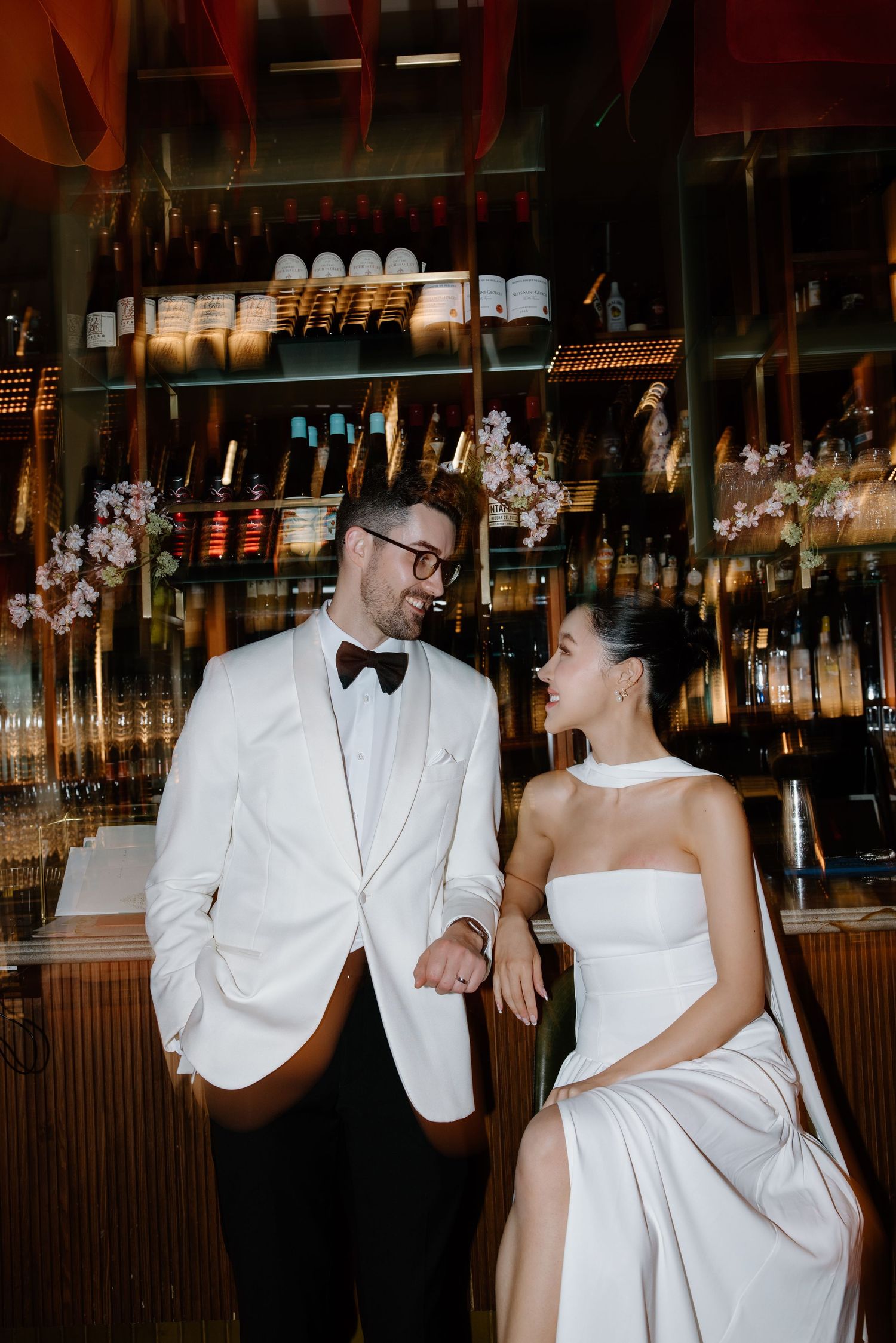 Elegant couple share intimate moment at upscale bar, wearing white formal attire against backdrop of liquor bottles.