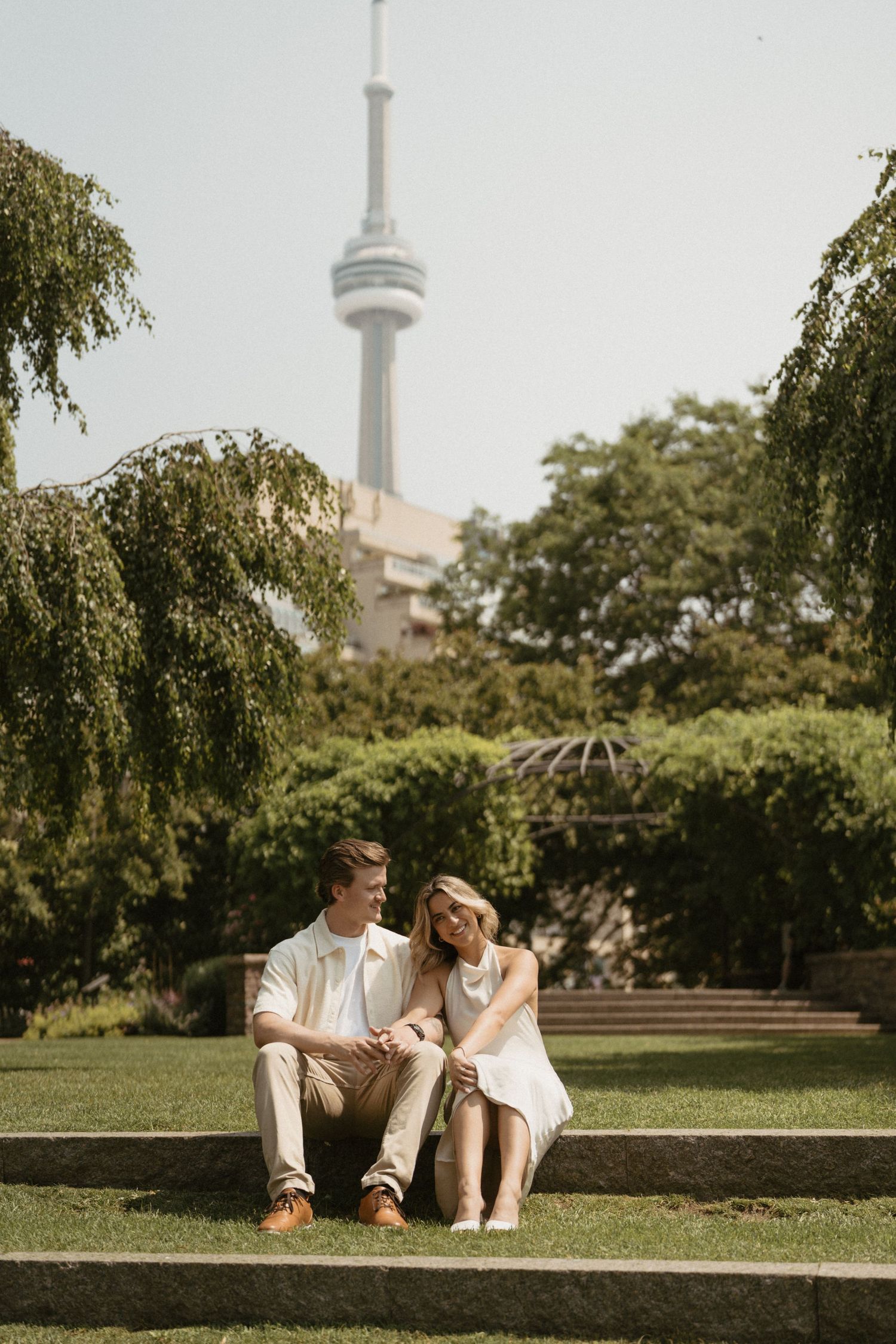 A couple sits together on stone steps with Toronto's CN Tower visible in the background on a sunny summer day.
