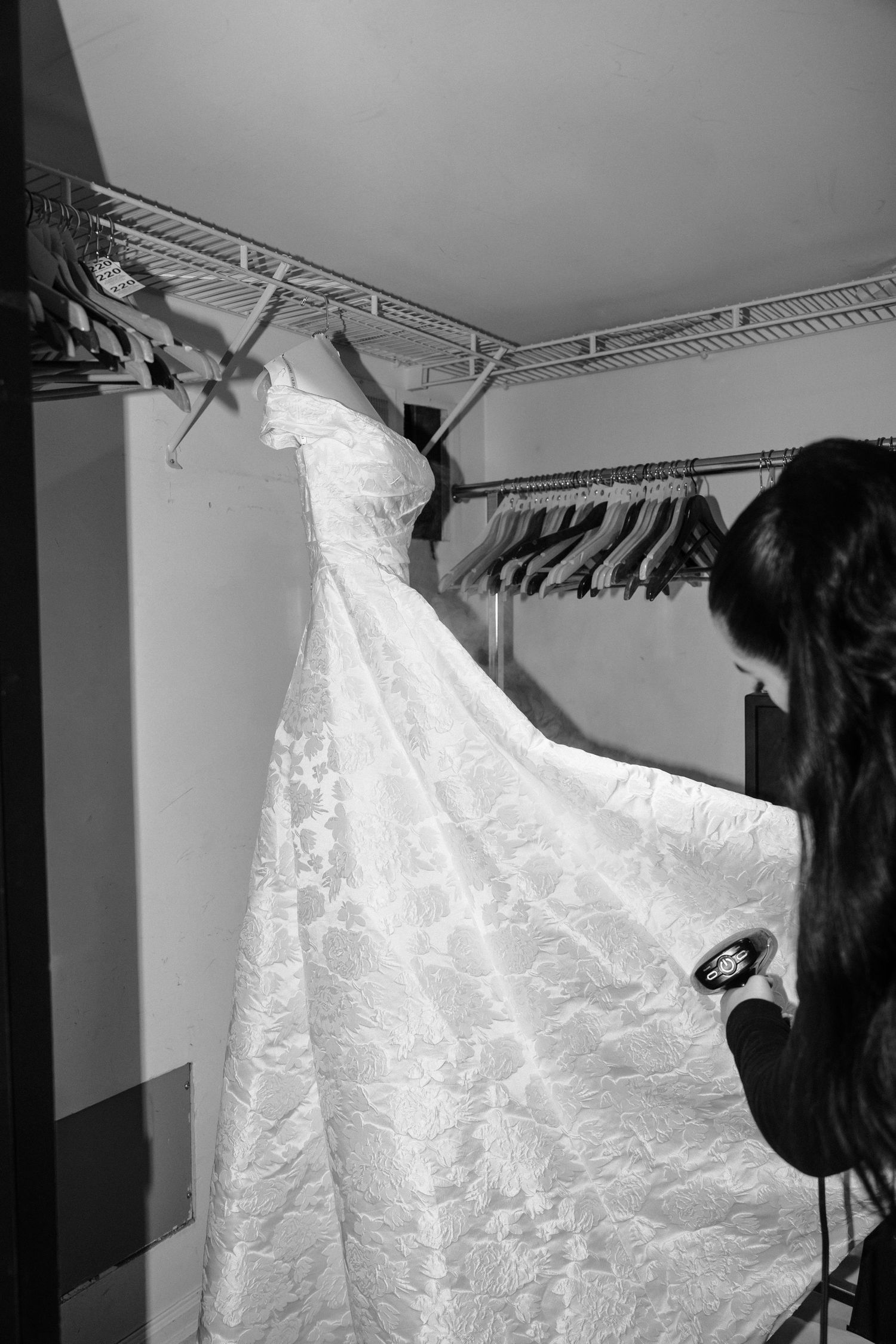 A white lace wedding dress with a long train hangs on a clothing rack in a dressing room.