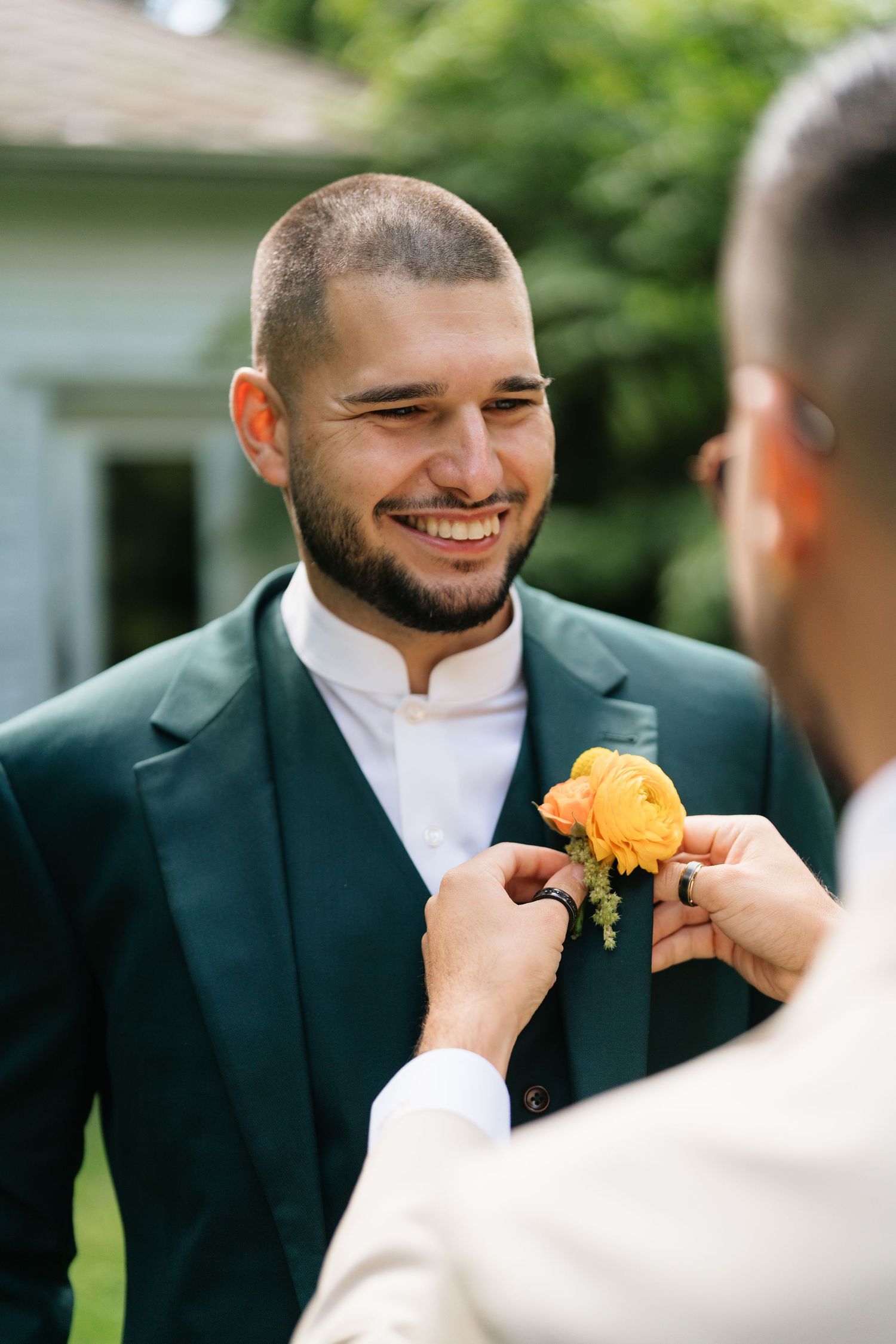A groomsman in a dark suit and white shirt smiles while having his tie adjusted before a wedding ceremony.