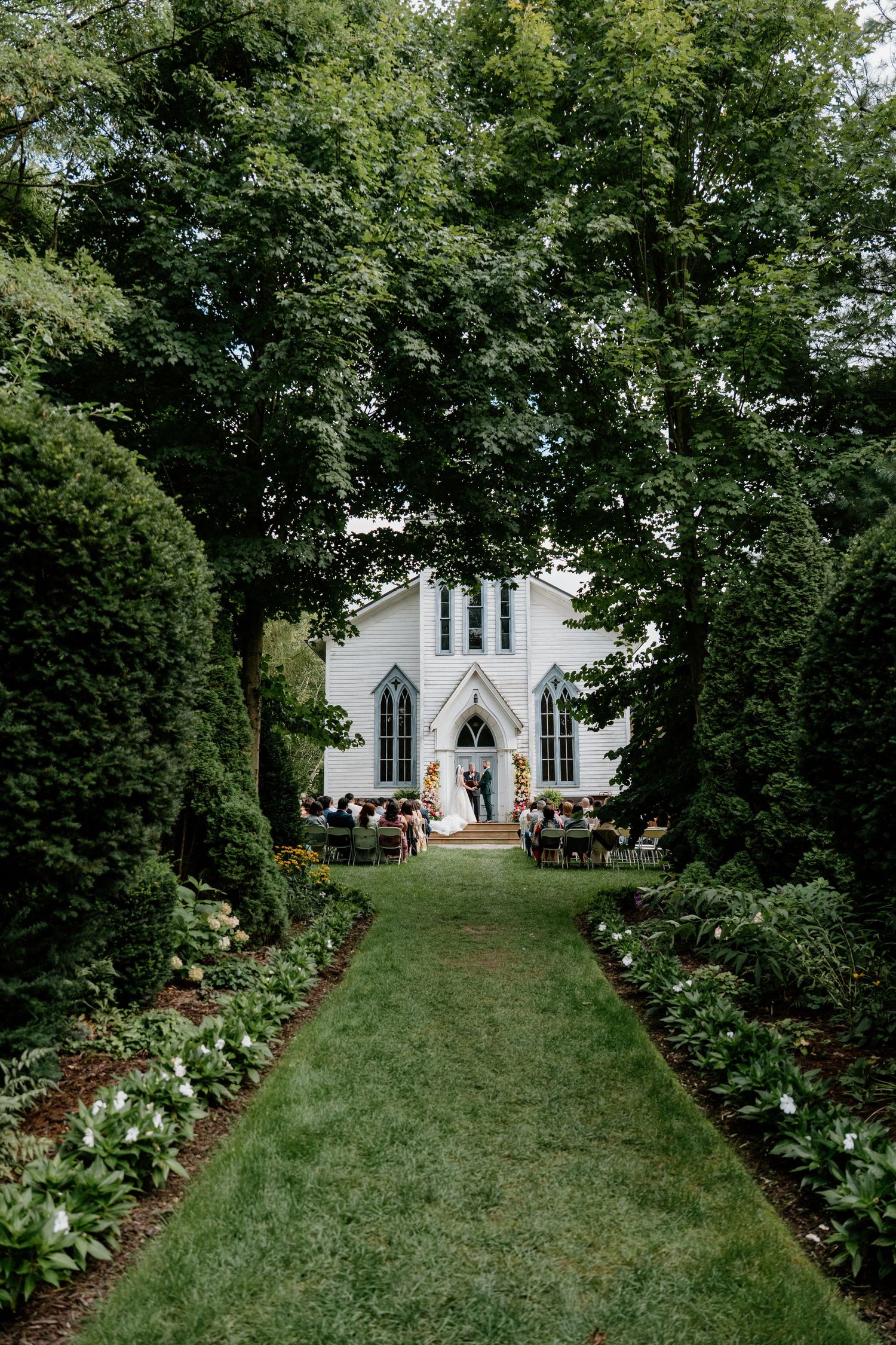A white chapel with Gothic windows sits at the end of a grassy aisle lined with flowering bushes in a garden setting.