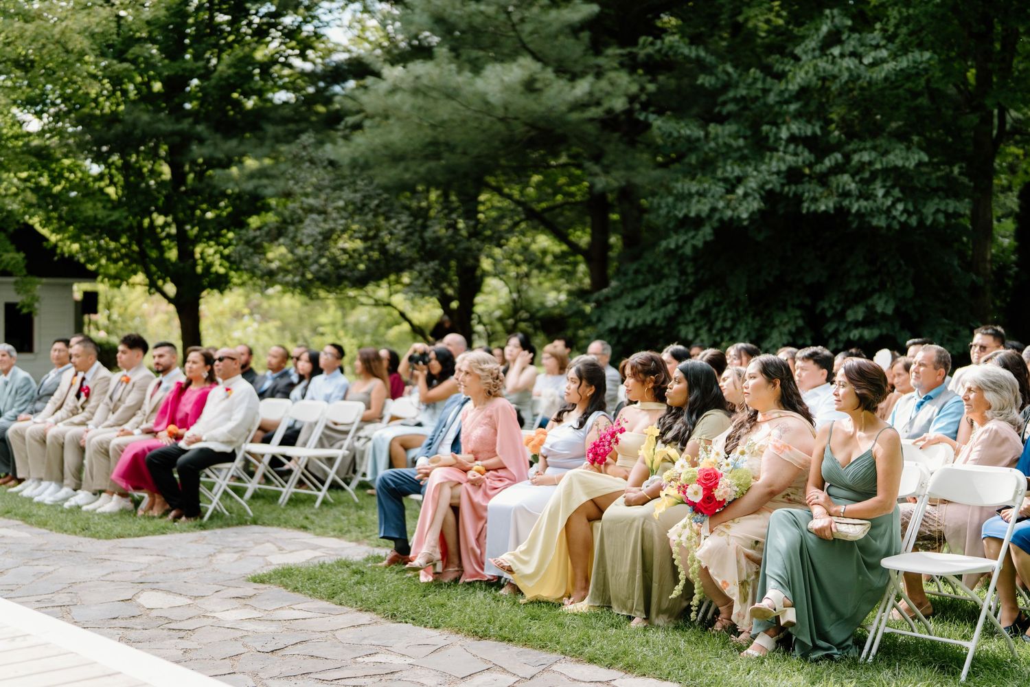 Wedding guests seated in rows of white chairs outdoors surrounded by lush green trees during a summer ceremony.