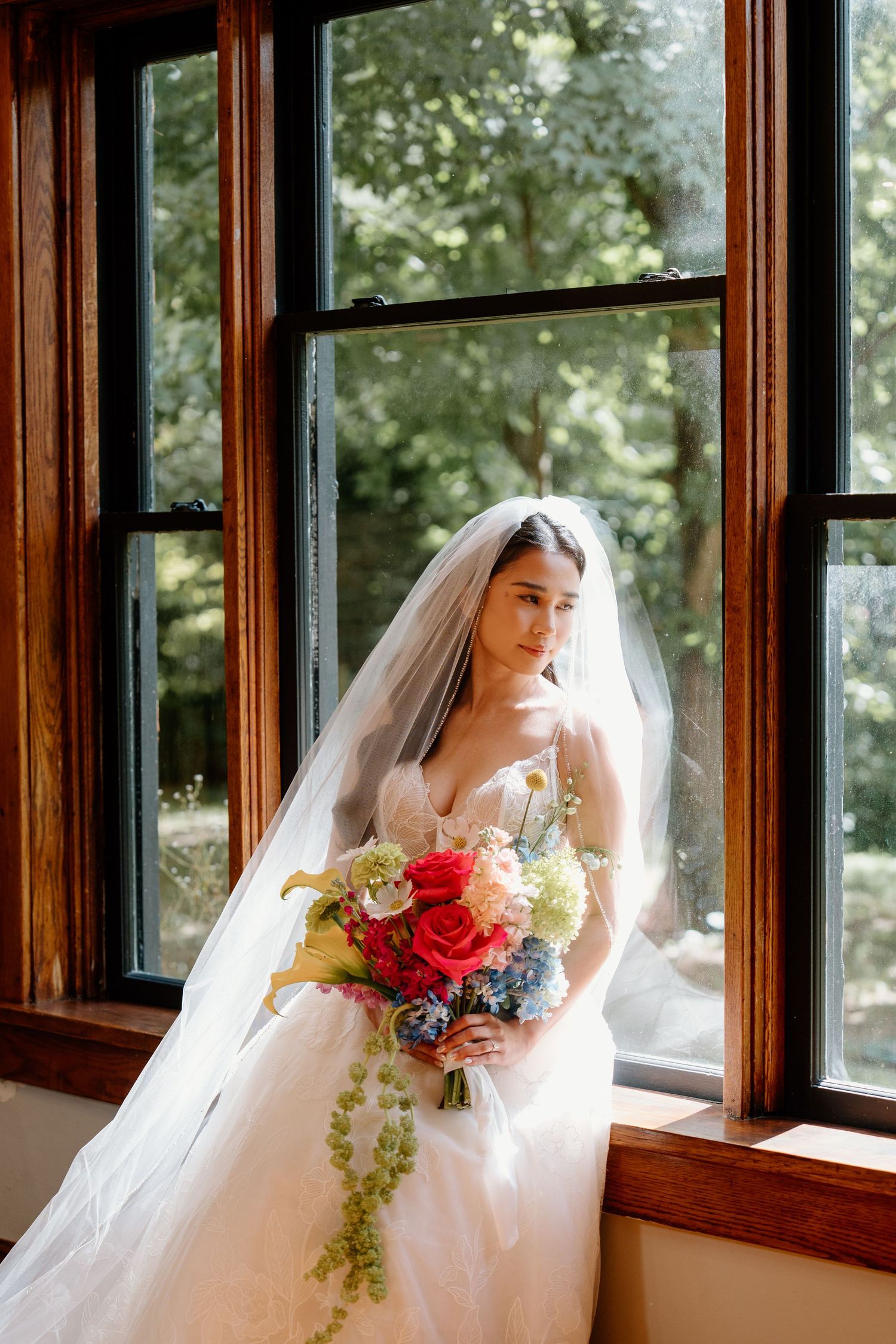A bride in an elegant white dress stands near wooden-framed windows holding a colorful bouquet with cascading greenery.