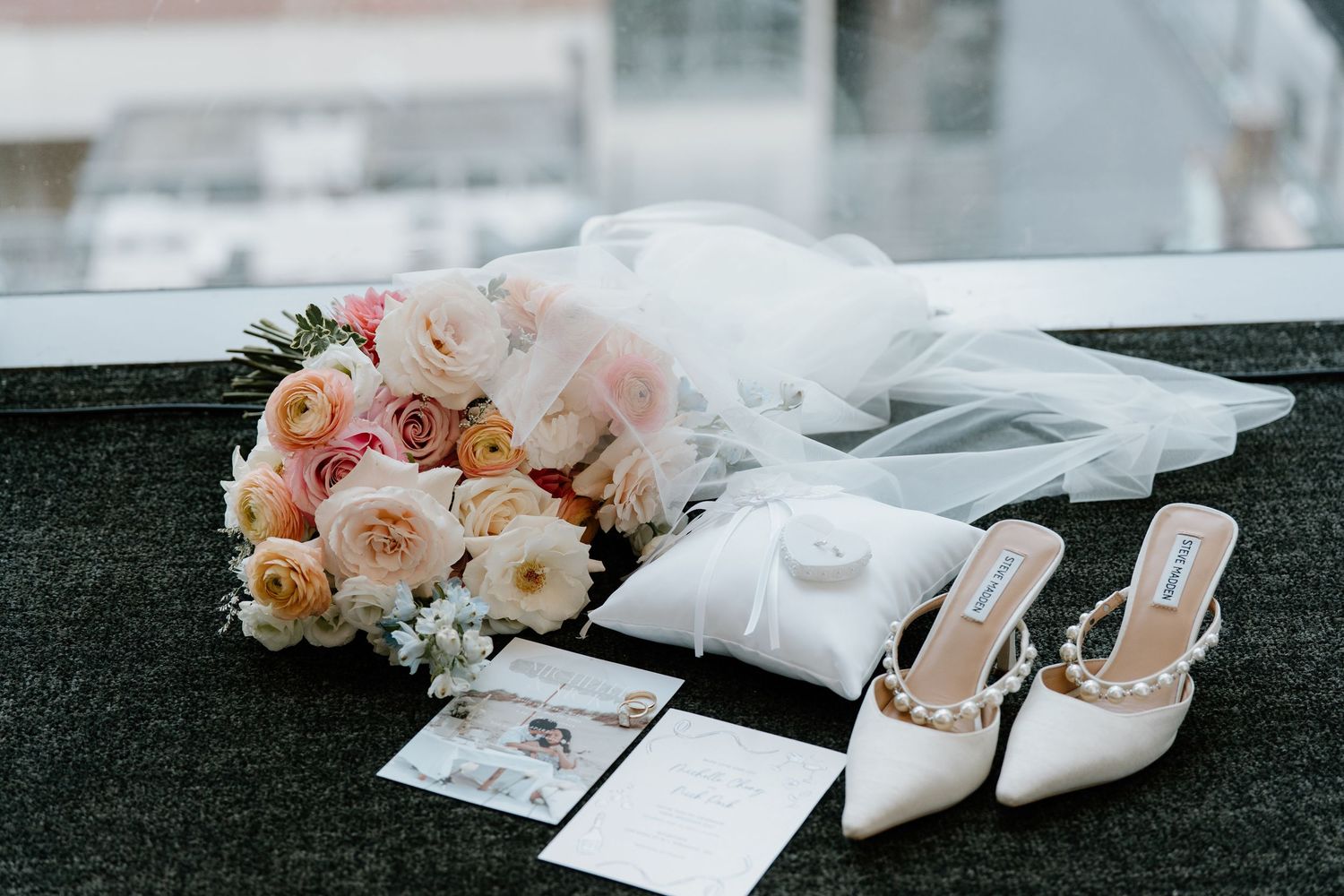 Bridal accessories including pink and peach roses, white veil, invitation cards and elegant white heeled mules on dark surface.