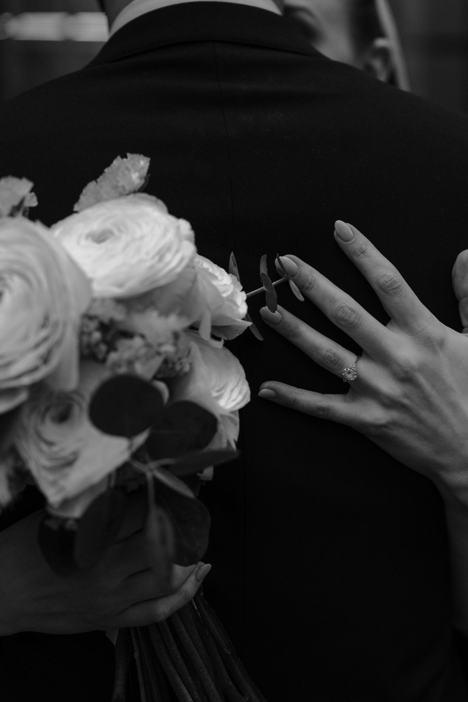 A hand wearing a wedding ring holds a bouquet of white roses against a black background in a monochrome photograph.