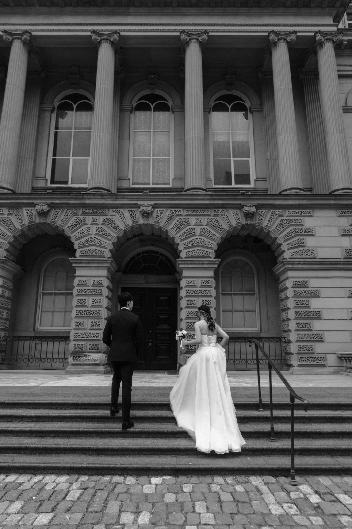 Black and white photo of a couple in wedding attire on the steps of a grand historic building with neoclassical columns.