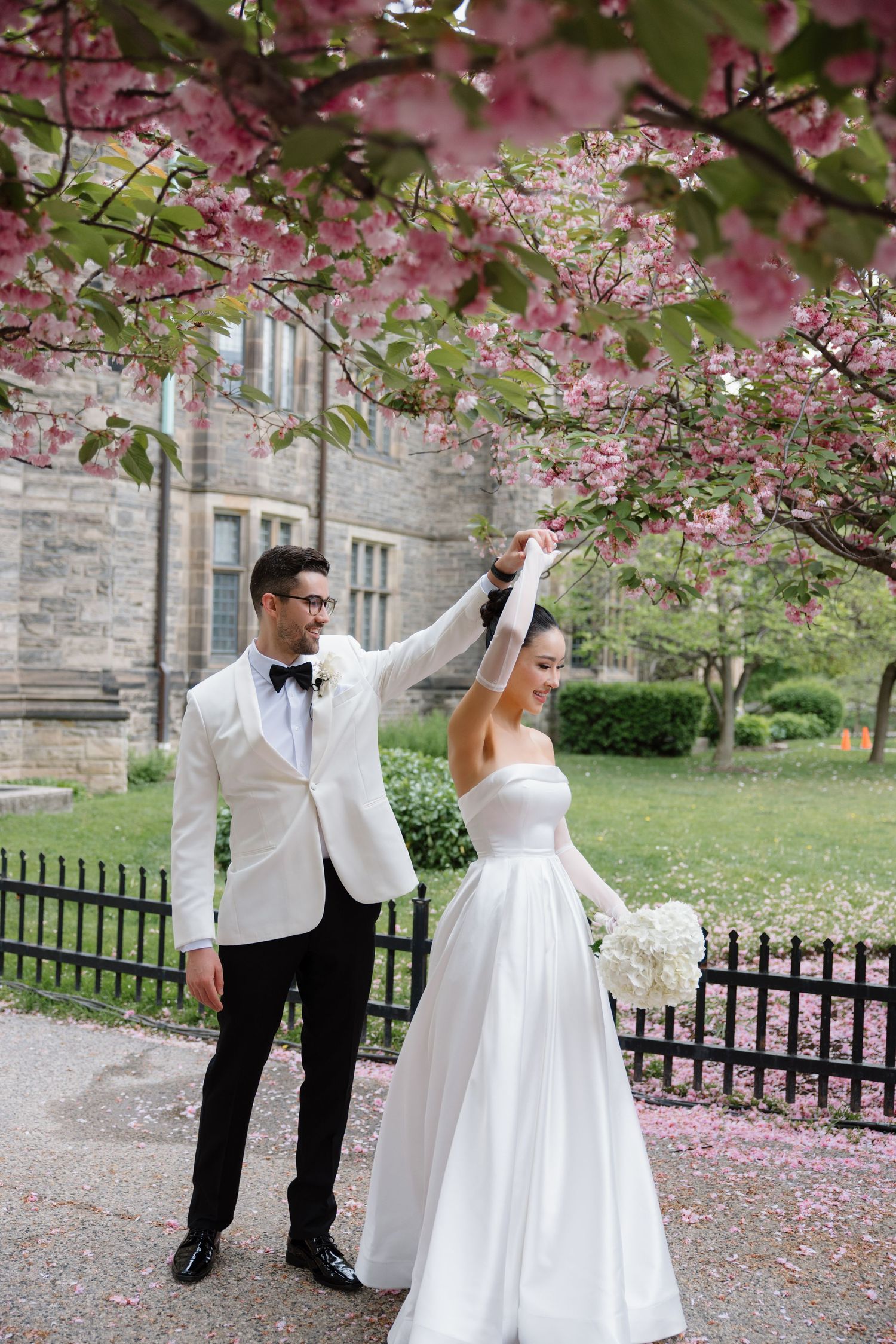 A couple in wedding attire stands beneath blooming pink cherry blossom trees on their special day.