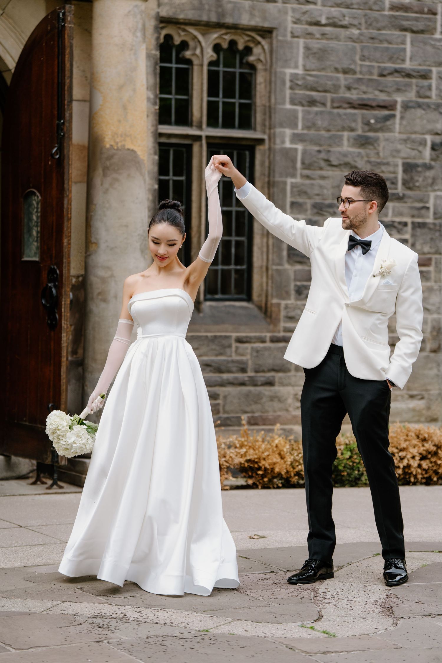 A couple in wedding attire performs a series of joyful dance moves outside a stone building.