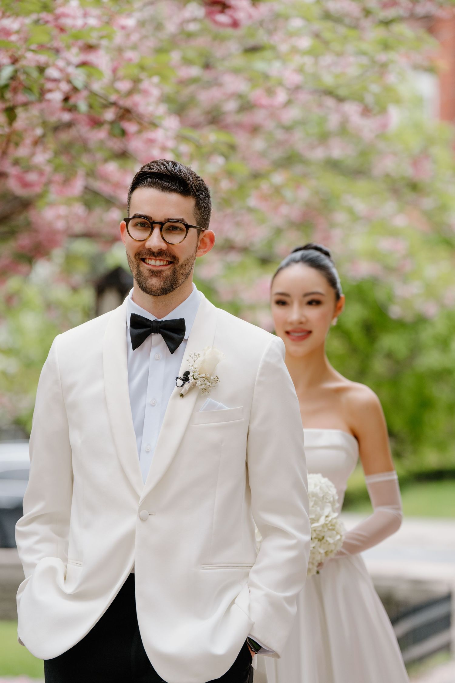 Newlywed couple poses outdoors with blooming cherry blossoms in the background during spring wedding.