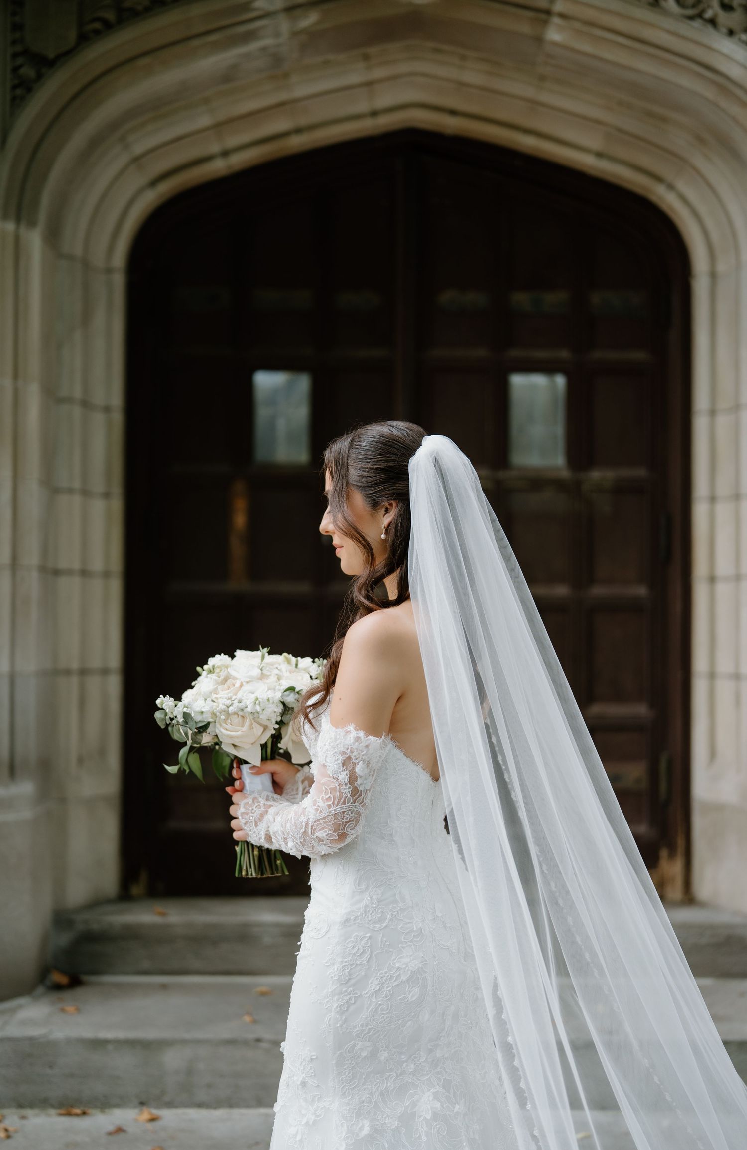 A bride in a white lace wedding gown with a flowing veil holds a white floral bouquet in front of a stone archway.