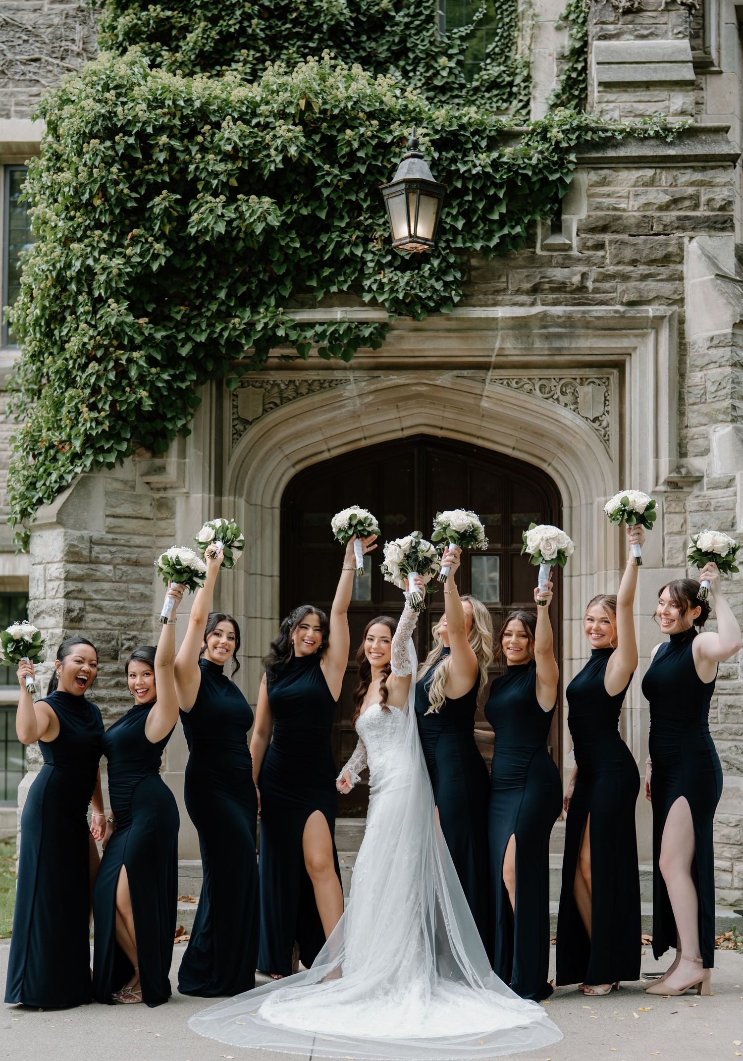 Wedding party in black dresses and white gowns celebrates joyfully outside a stone building with gothic architecture.