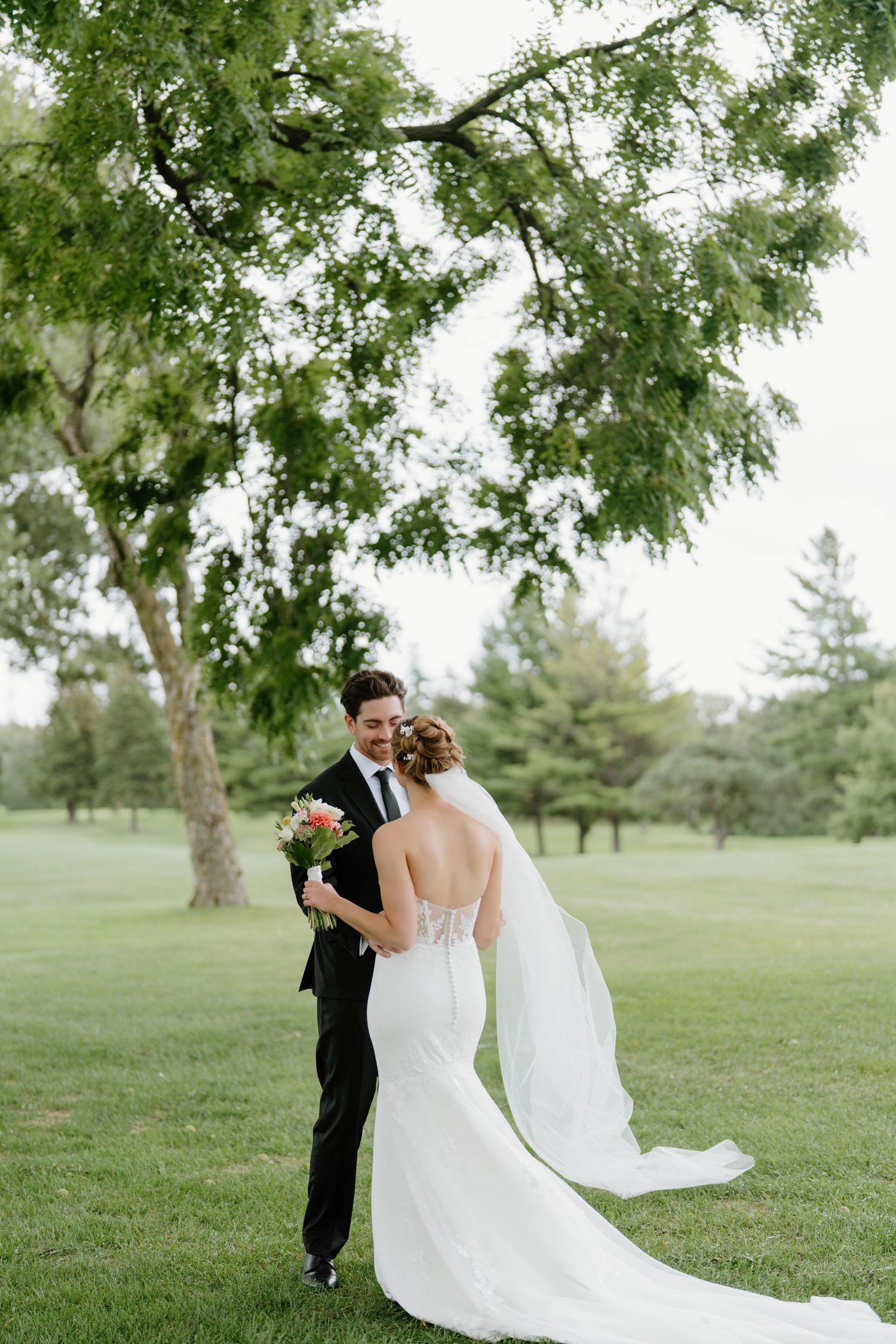 Couple in wedding attire walking together on a lush green lawn with trees in the background.
