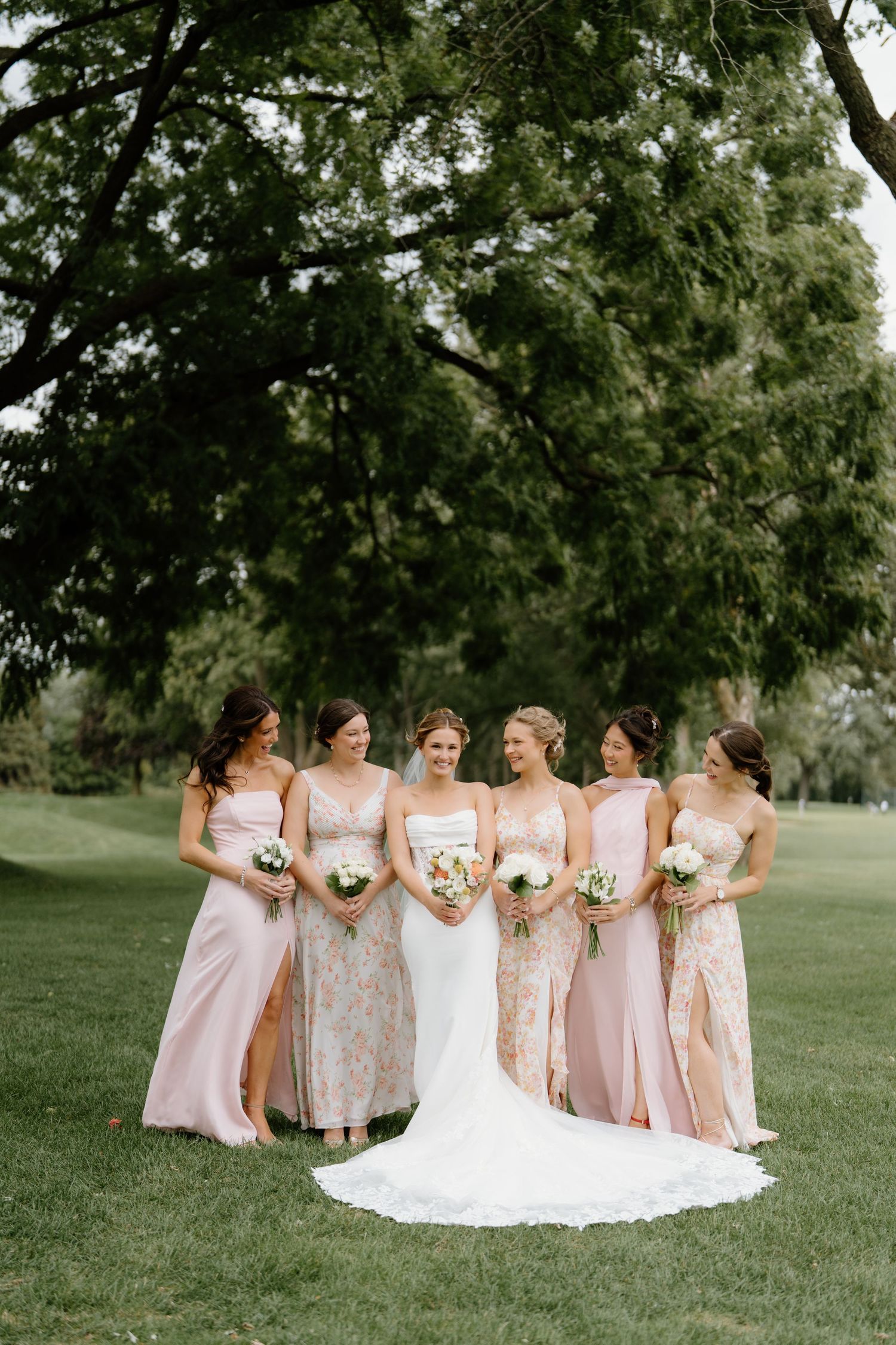 Bridesmaids in soft pink dresses and floral patterns gather on a lush green lawn for elegant wedding photos.