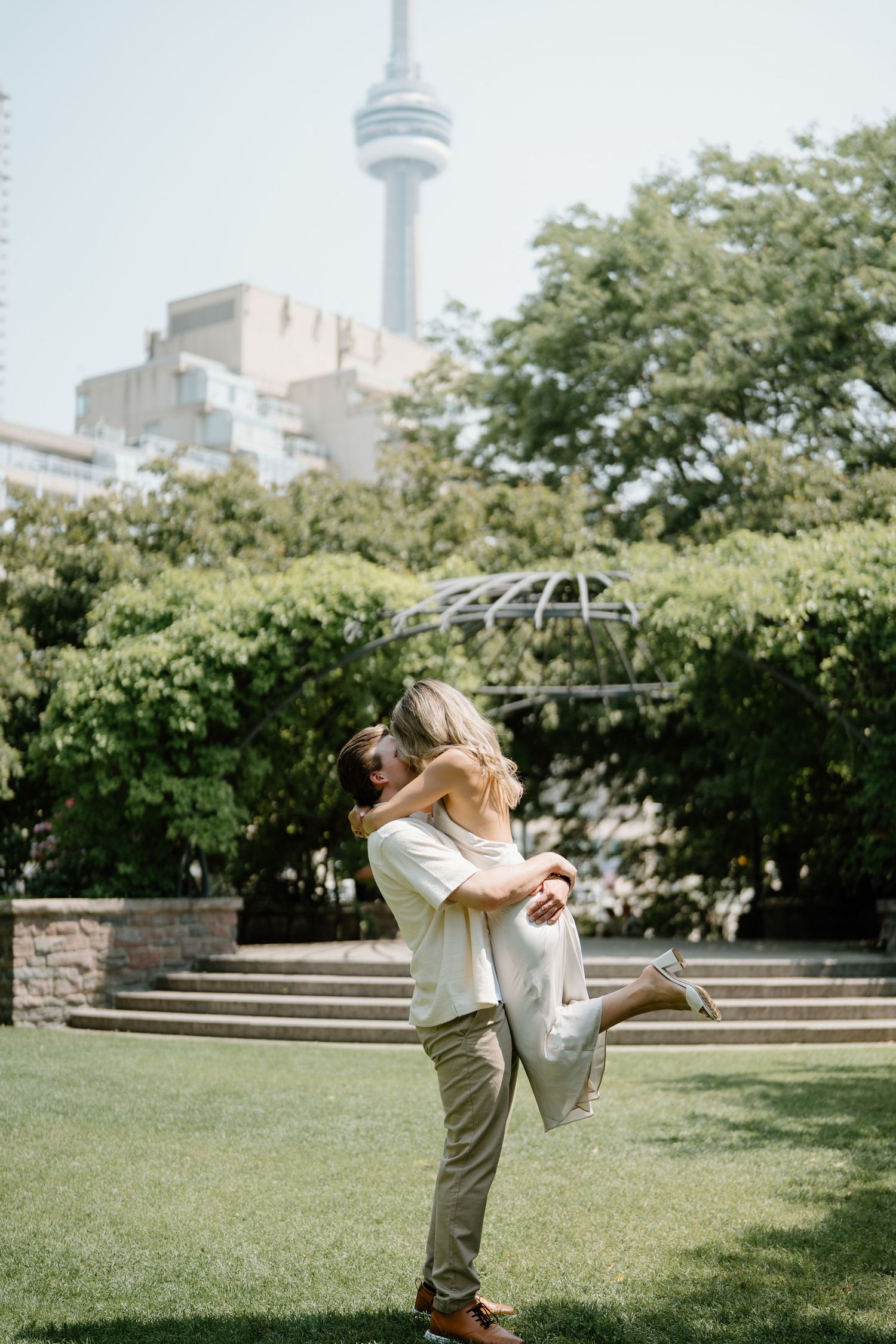 A couple shares a tender moment while dancing in a sunny urban park with city architecture in the background.