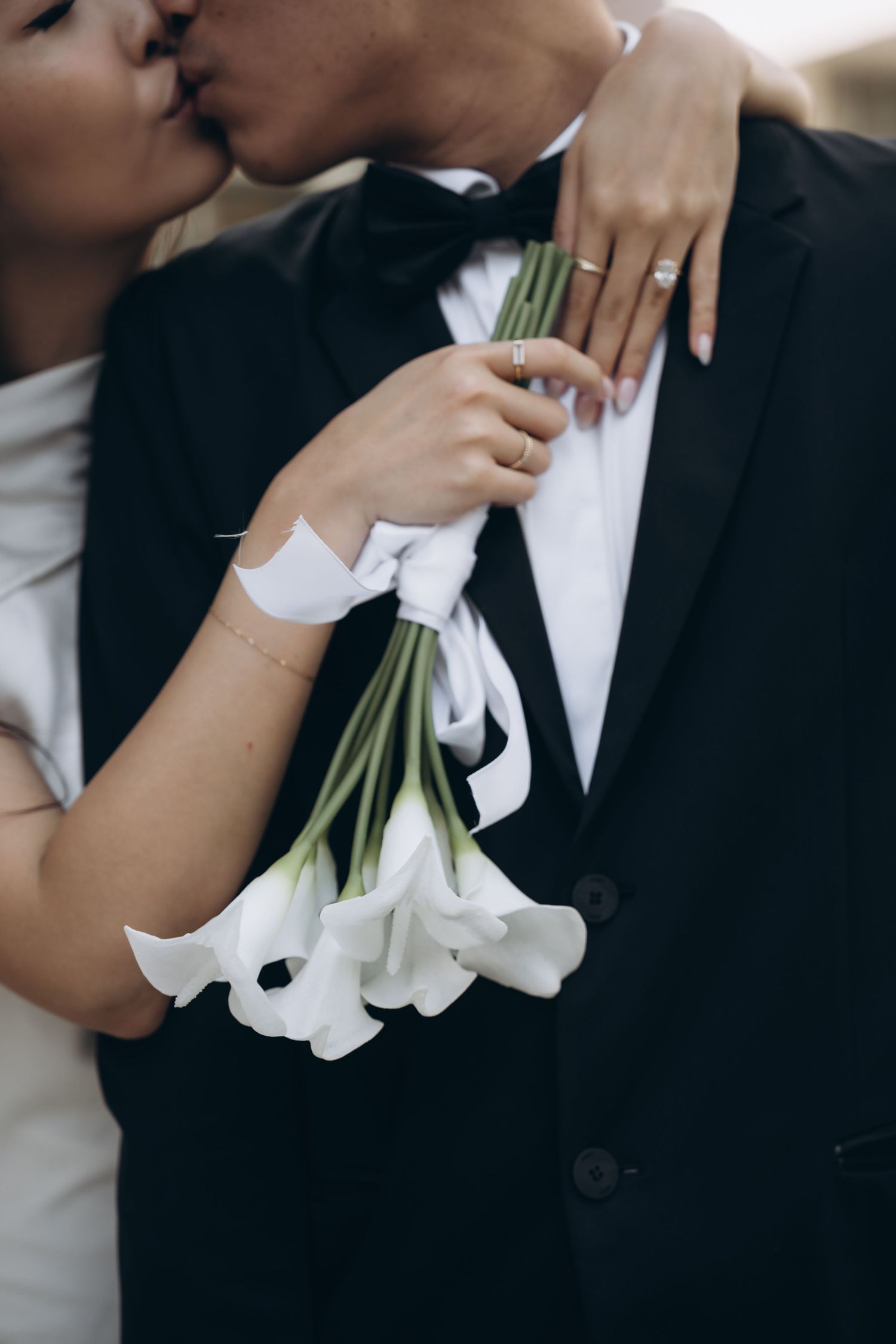 Elegant couple shares tender kiss while holding white calla lilies wrapped in ribbon against black tuxedo.