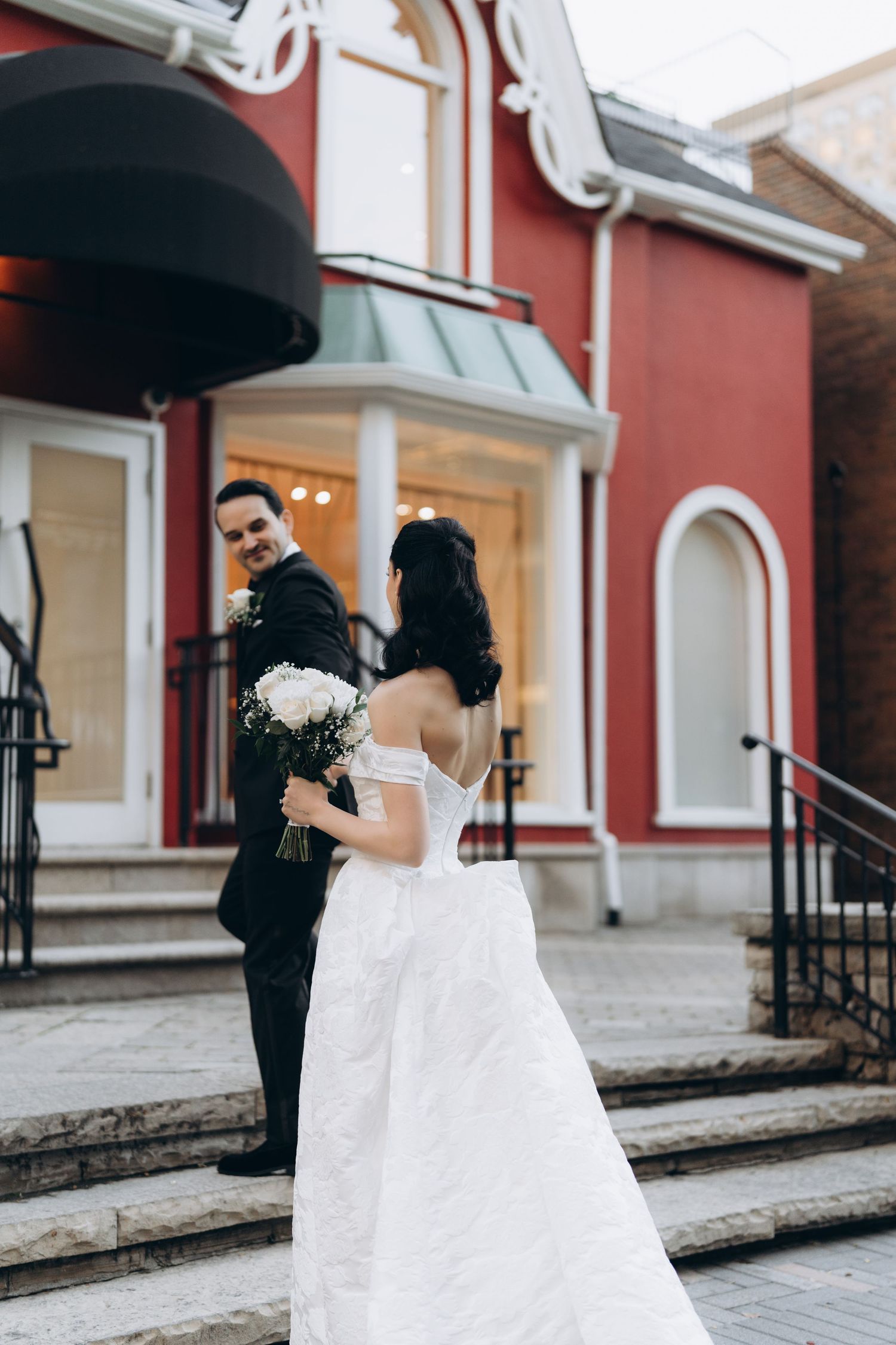 Wedding couple shares a playful dance moment in front of a red-painted building with white trim.