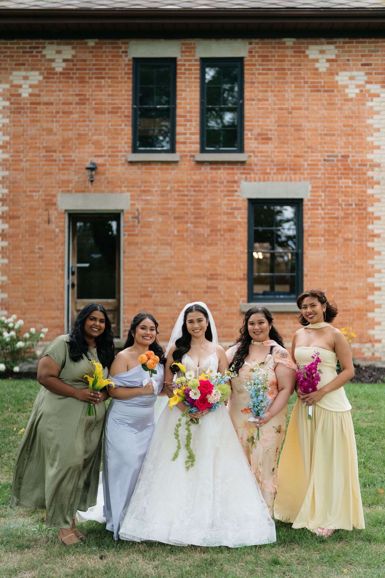Wedding party in pastel yellow dresses and olive suits sits casually on grass against brick building.