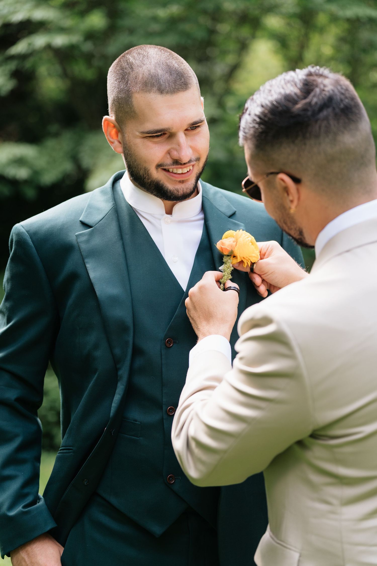 Two men in formal attire share a joyful moment as one pins a yellow boutonniere on the other's lapel.