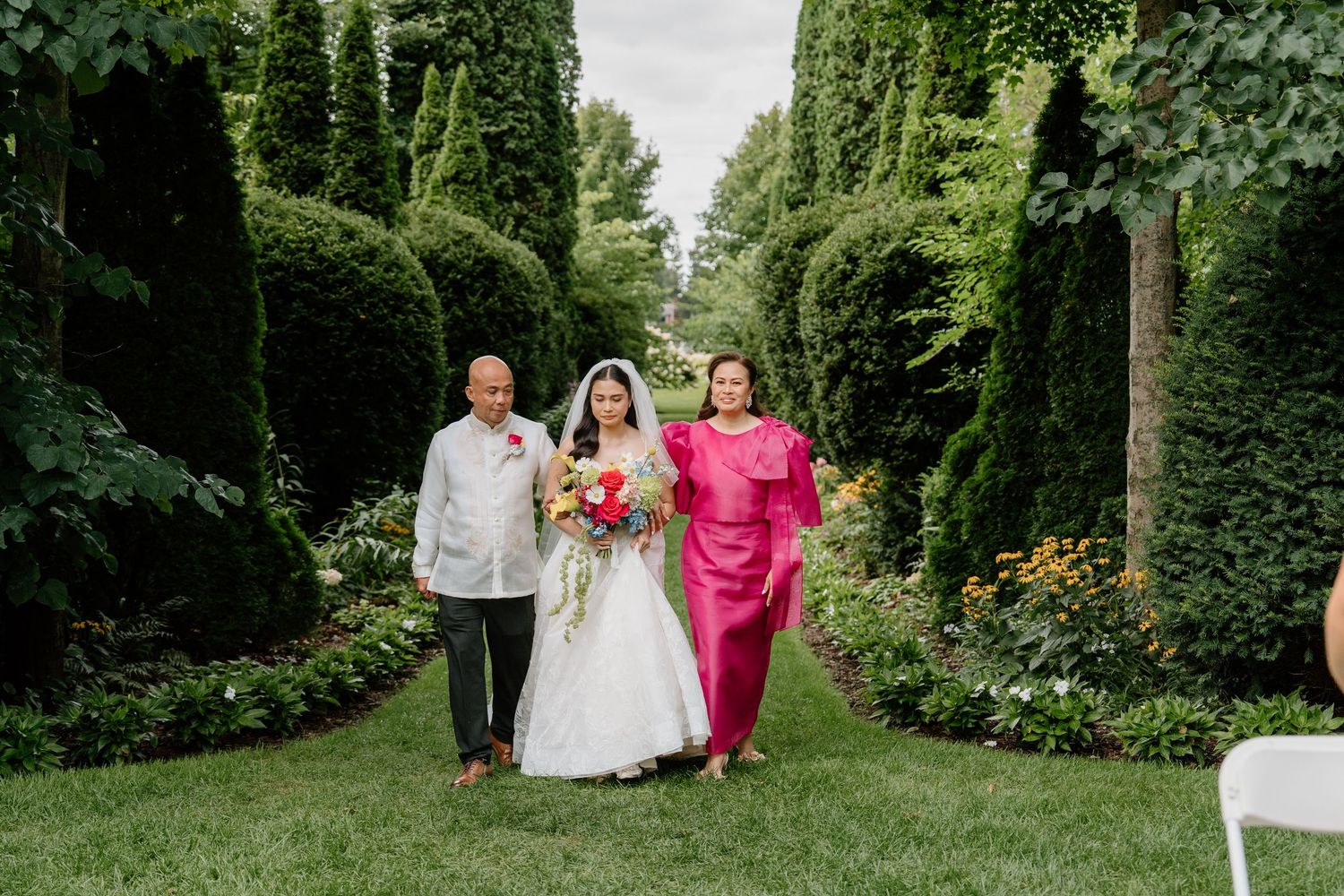 Wedding procession walks through a lush garden path with tall hedges and greenery.