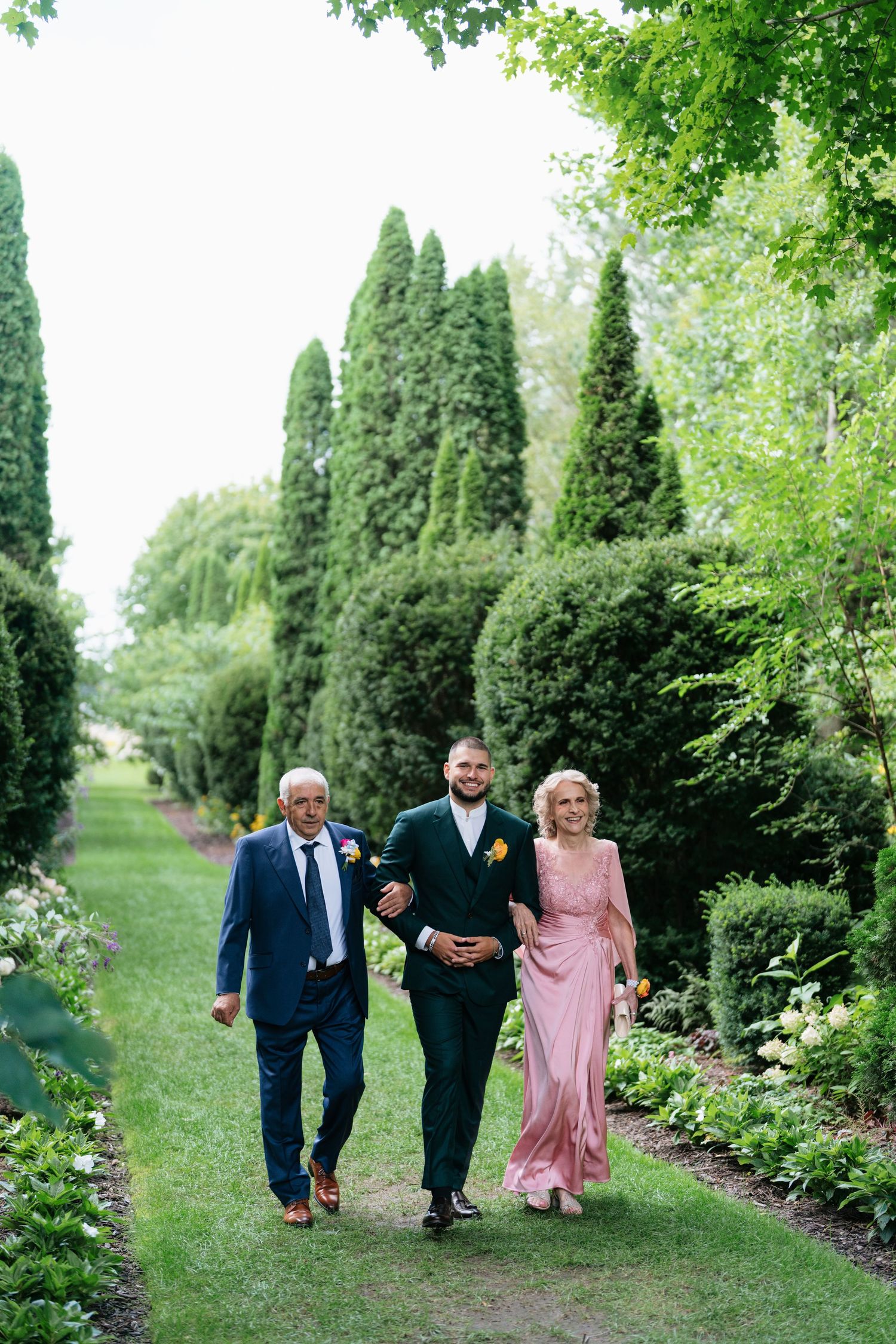 Three people walk together on a lush garden path lined with tall evergreen trees and flowering plants.