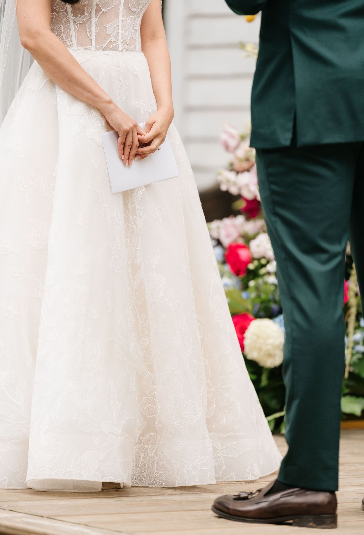 Detail shot of wedding dress and emerald green suit at outdoor ceremony with red floral decorations.