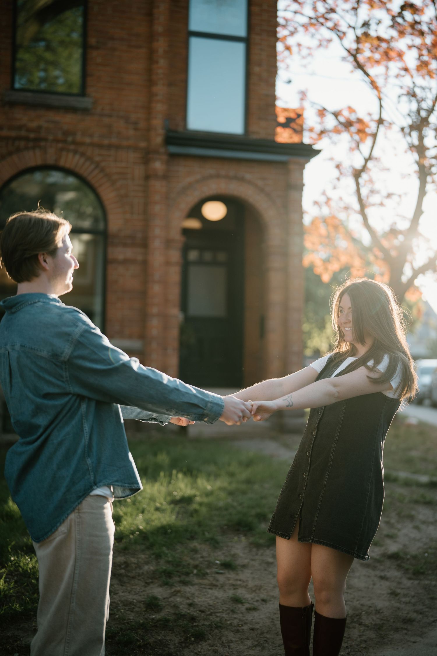 A romantic couple holding hands while dancing outside a brick building during autumn sunset.