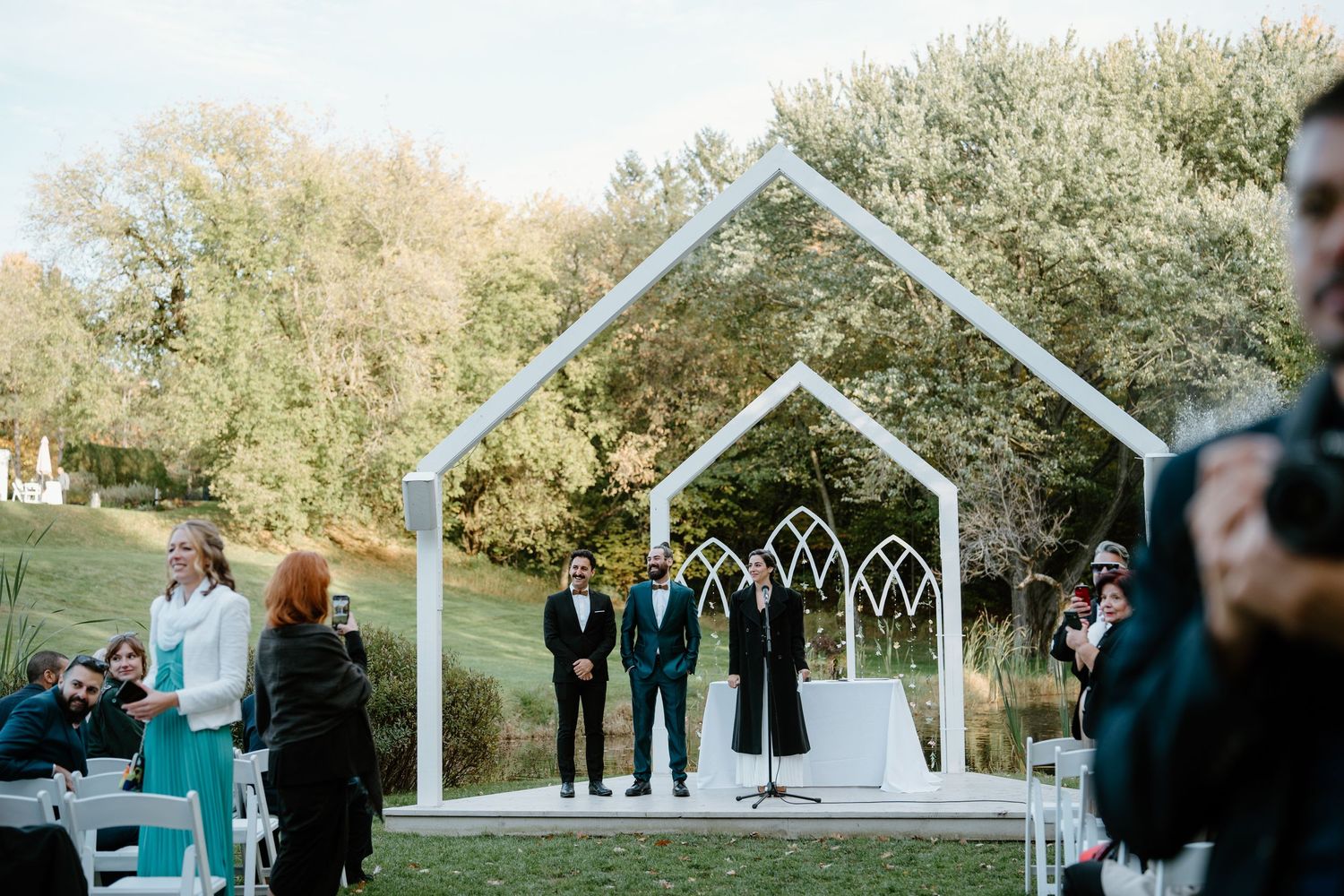 An outdoor wedding ceremony under a white architectural arch with guests seated on white chairs.