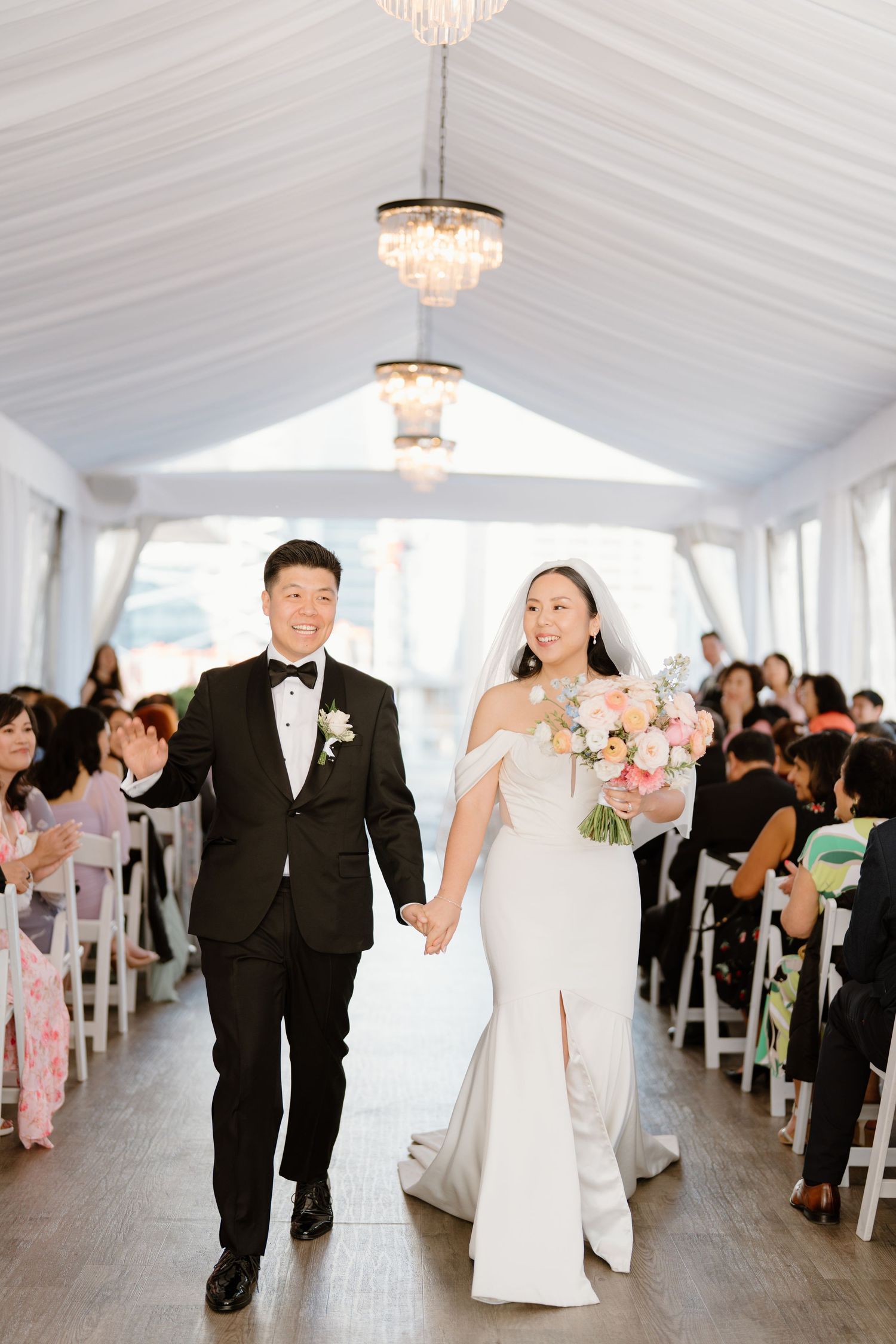 A wedding ceremony takes place indoors under crystal chandeliers with guests seated in white chairs along the aisle.