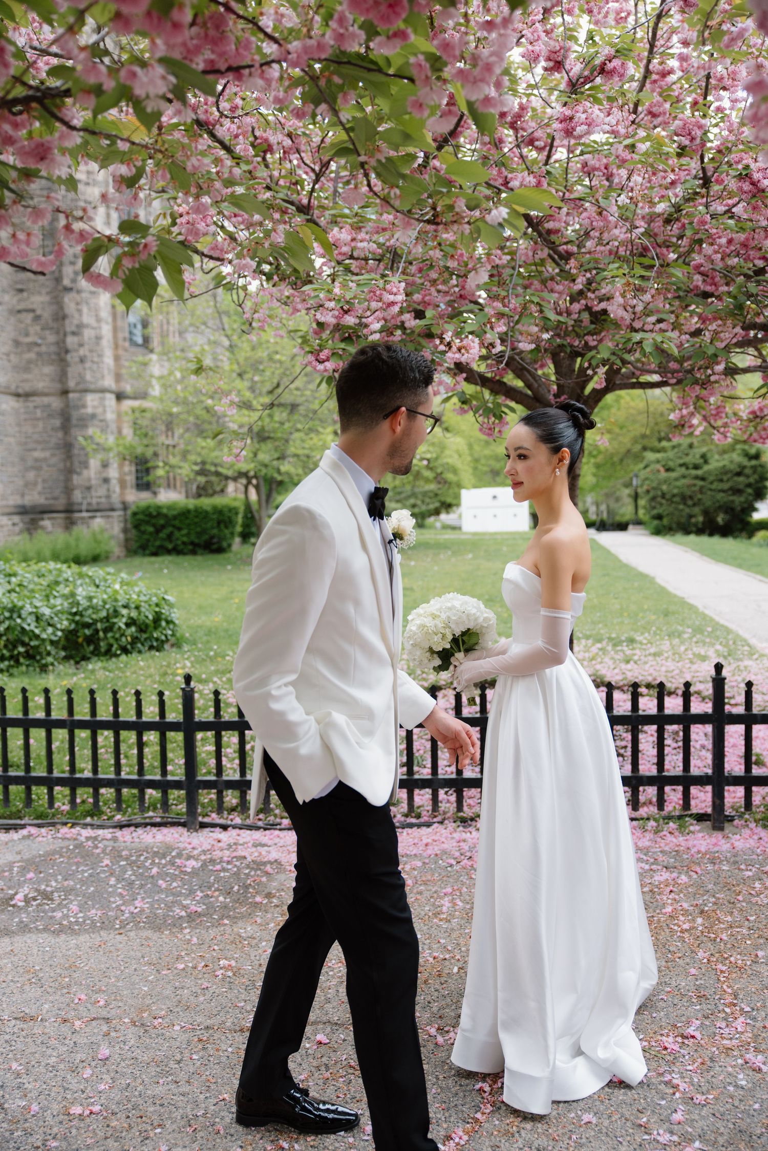 Couple stands beneath blooming cherry blossoms with bride in white gown and groom in white jacket and black pants.