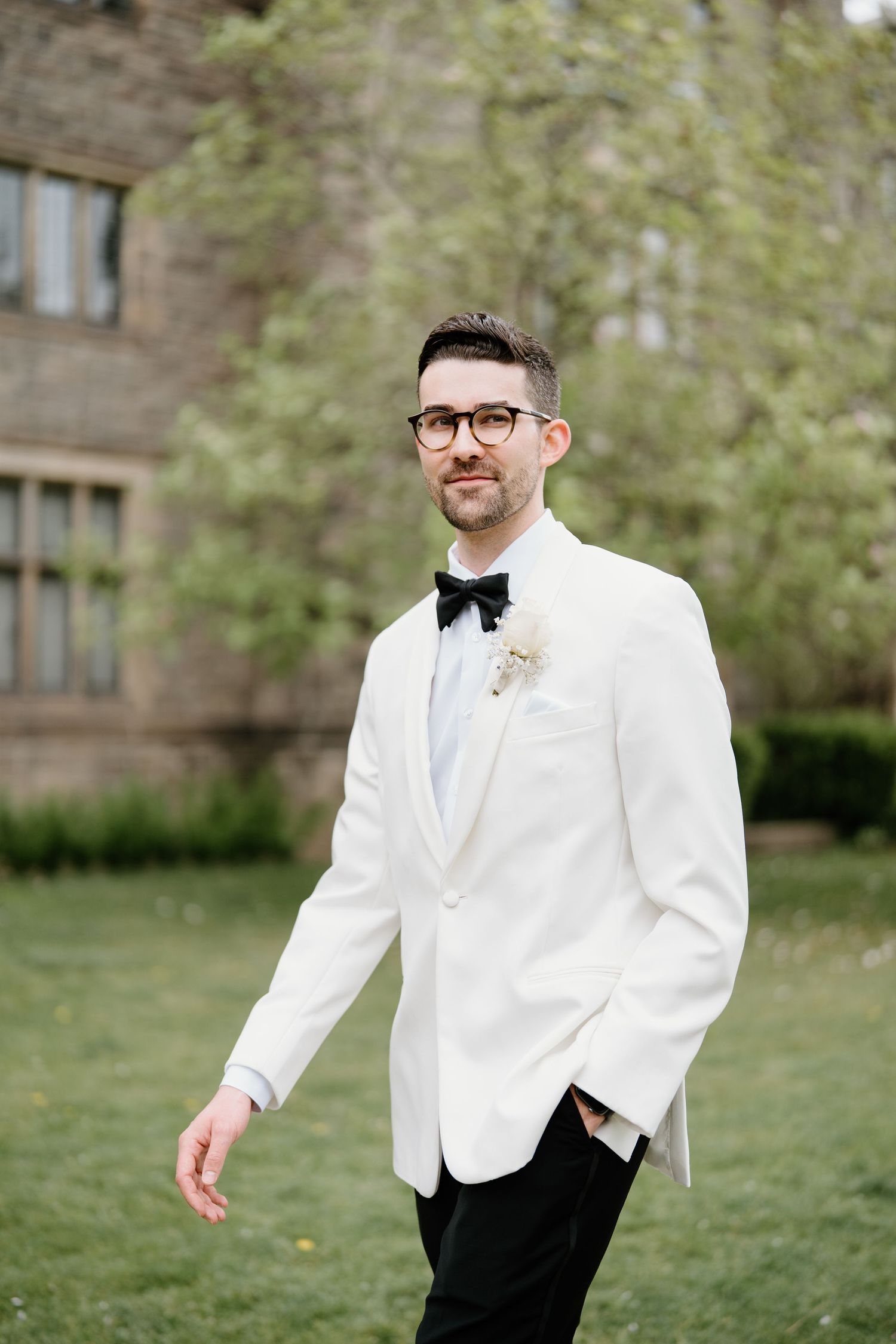 Groom in white tuxedo jacket and black bow tie poses in garden setting.