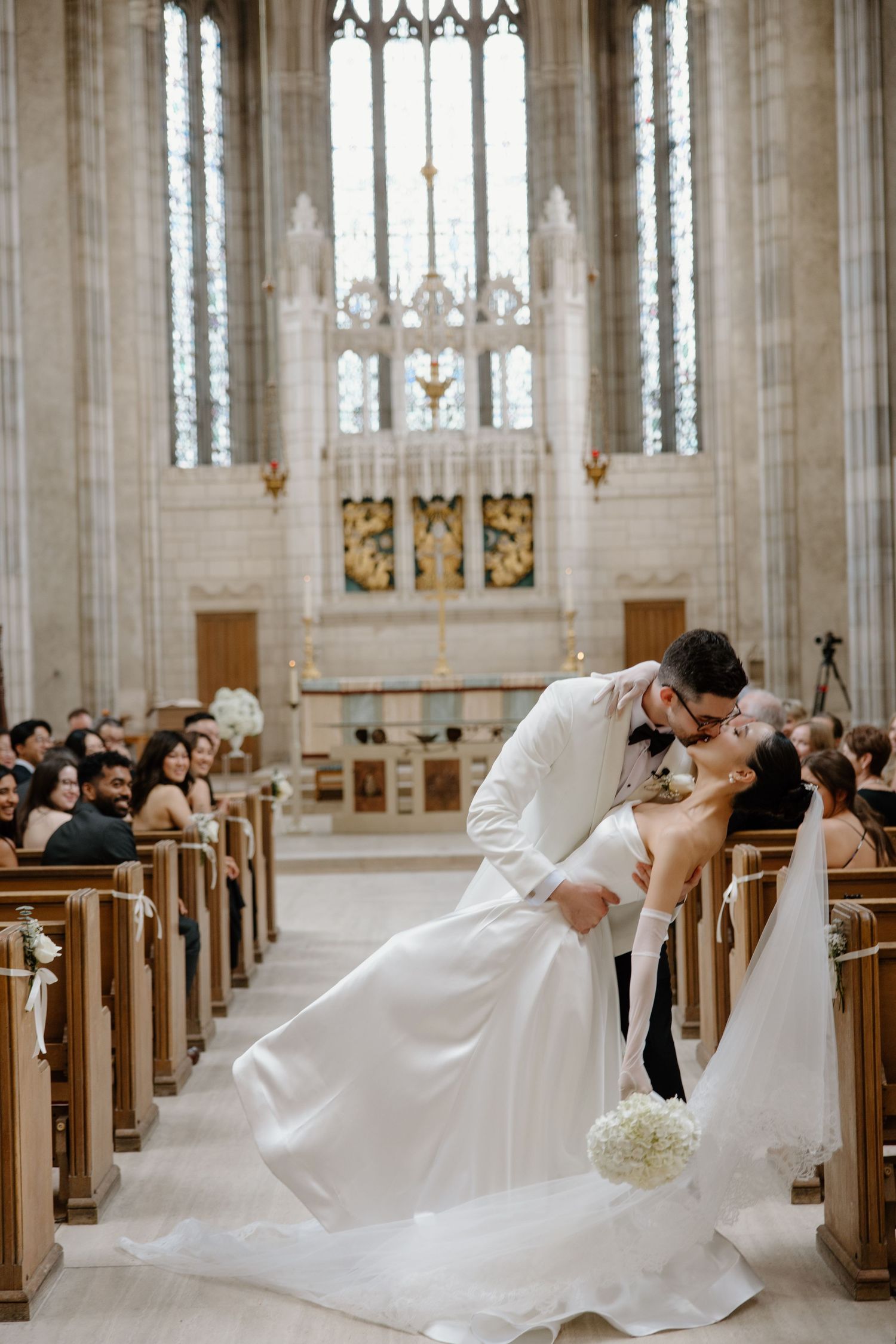 A couple shares a romantic dip kiss during their wedding ceremony in a grand cathedral with stained glass windows and wooden pews.