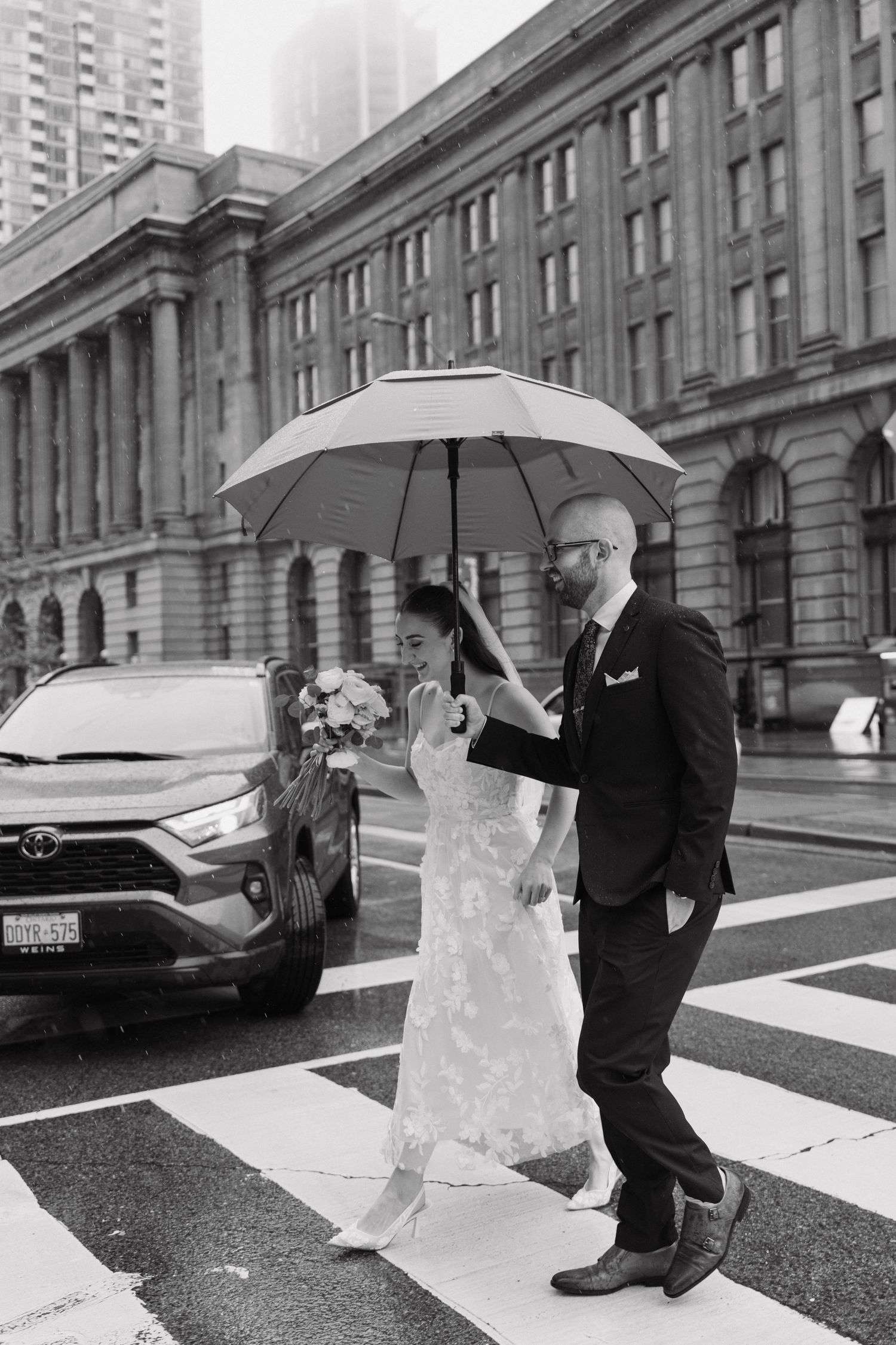 A couple shares a romantic moment under umbrellas in the rain on a city street during their wedding photoshoot.