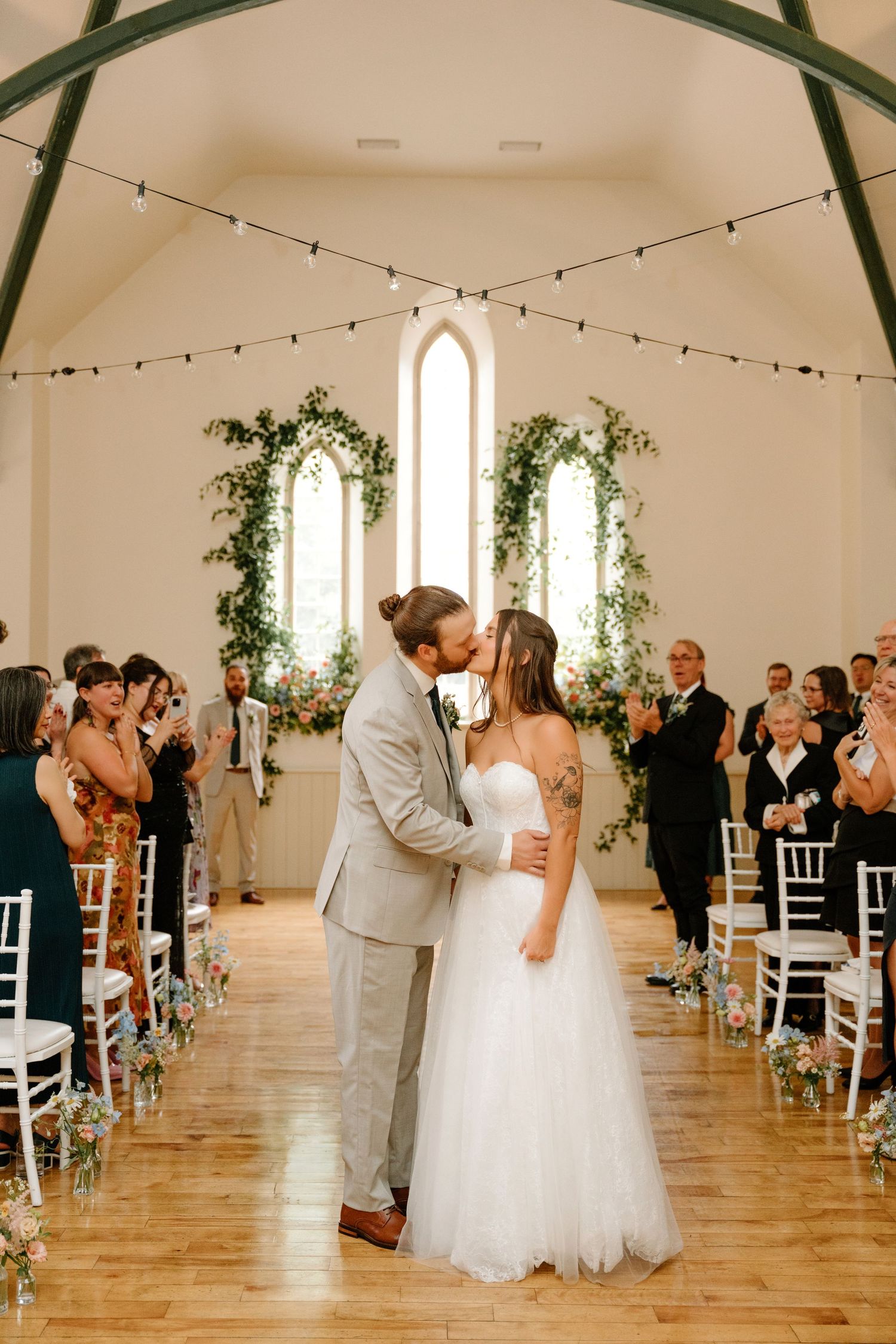 A romantic wedding ceremony moment in a bright white chapel with string lights and greenery decorations.