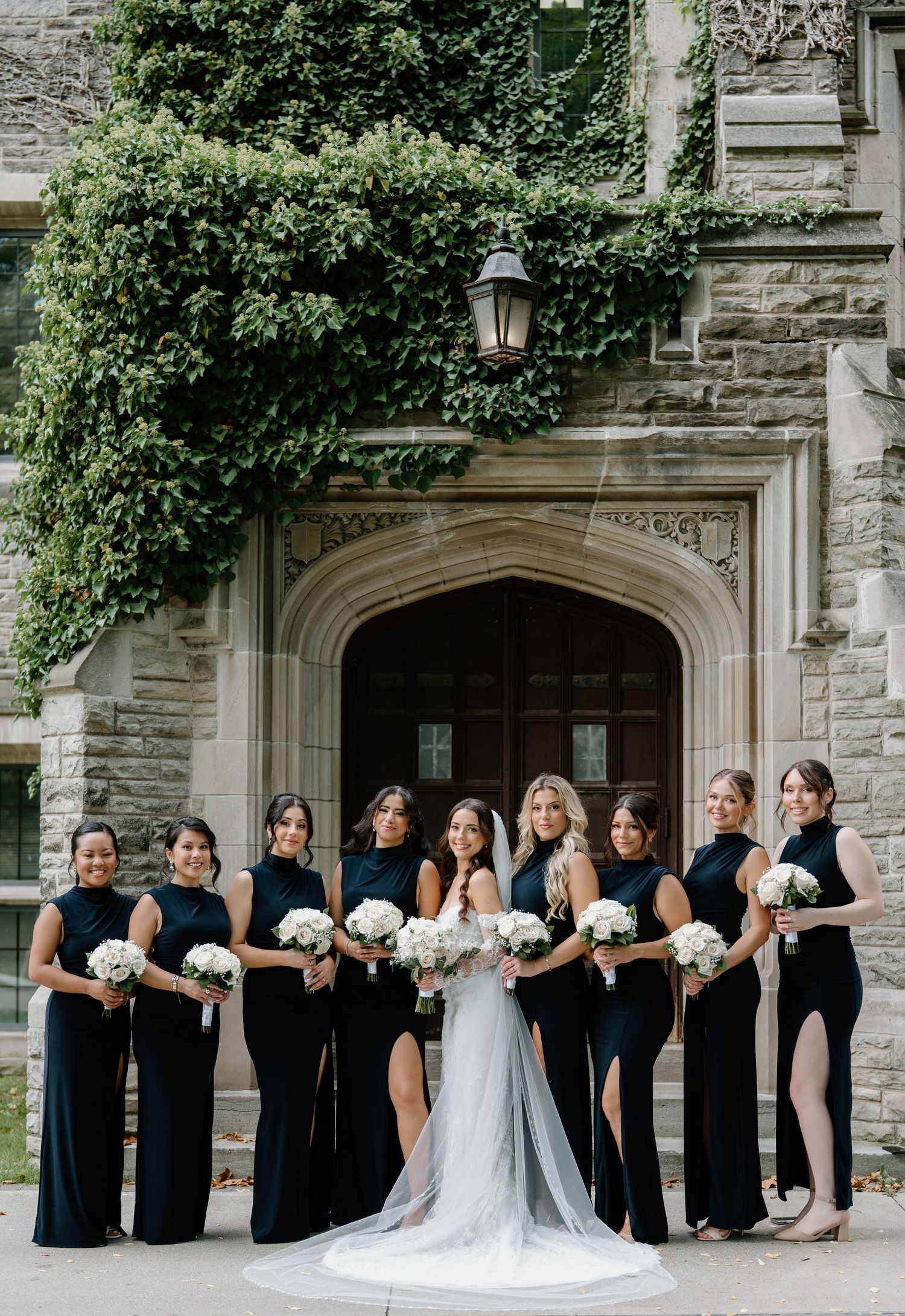 Elegant bridal party in navy blue dresses and white bouquets poses in front of gothic stone archway with climbing ivy.