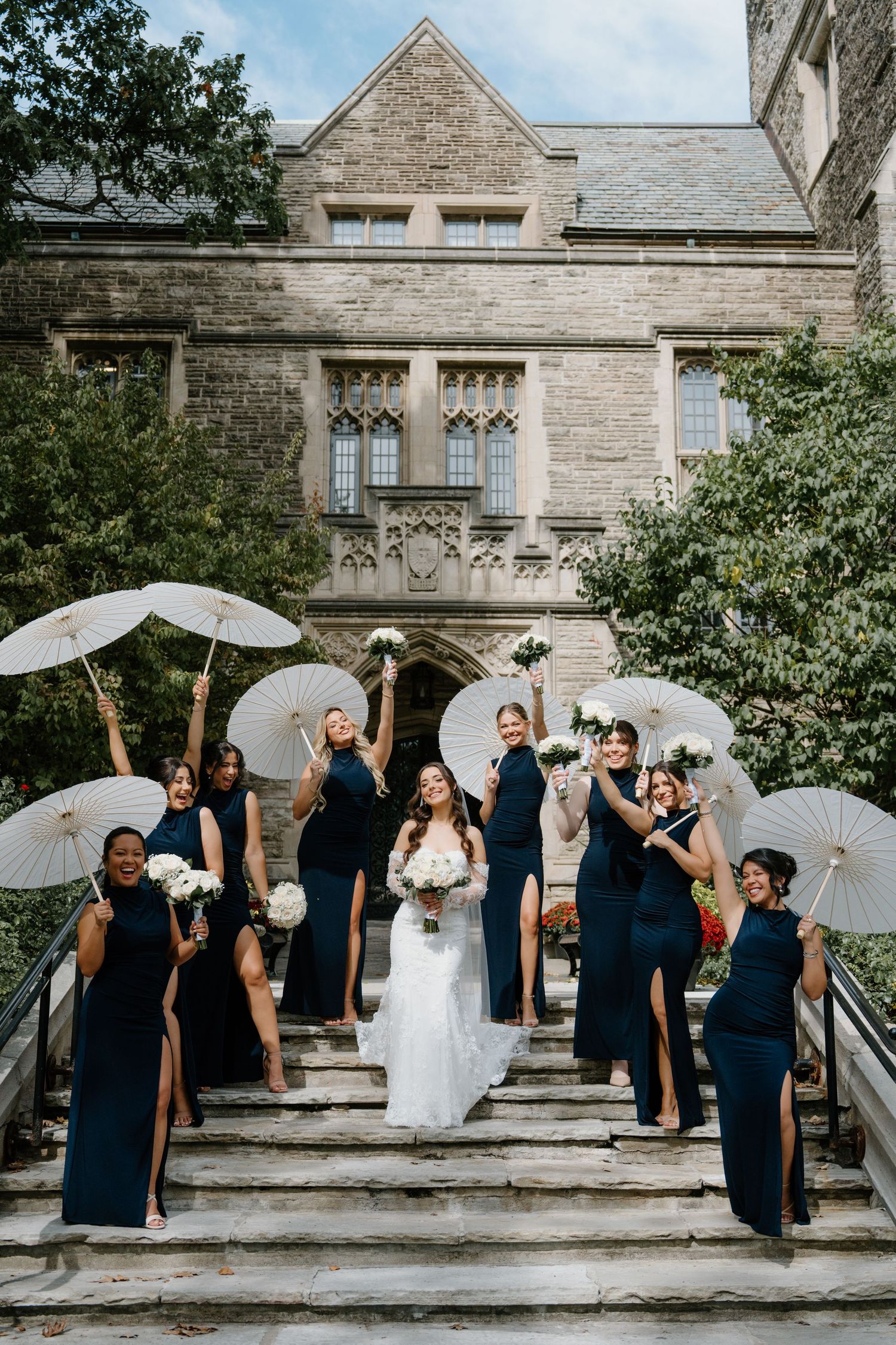 Wedding party with navy blue dresses and white parasols pose on stone steps of historic building.