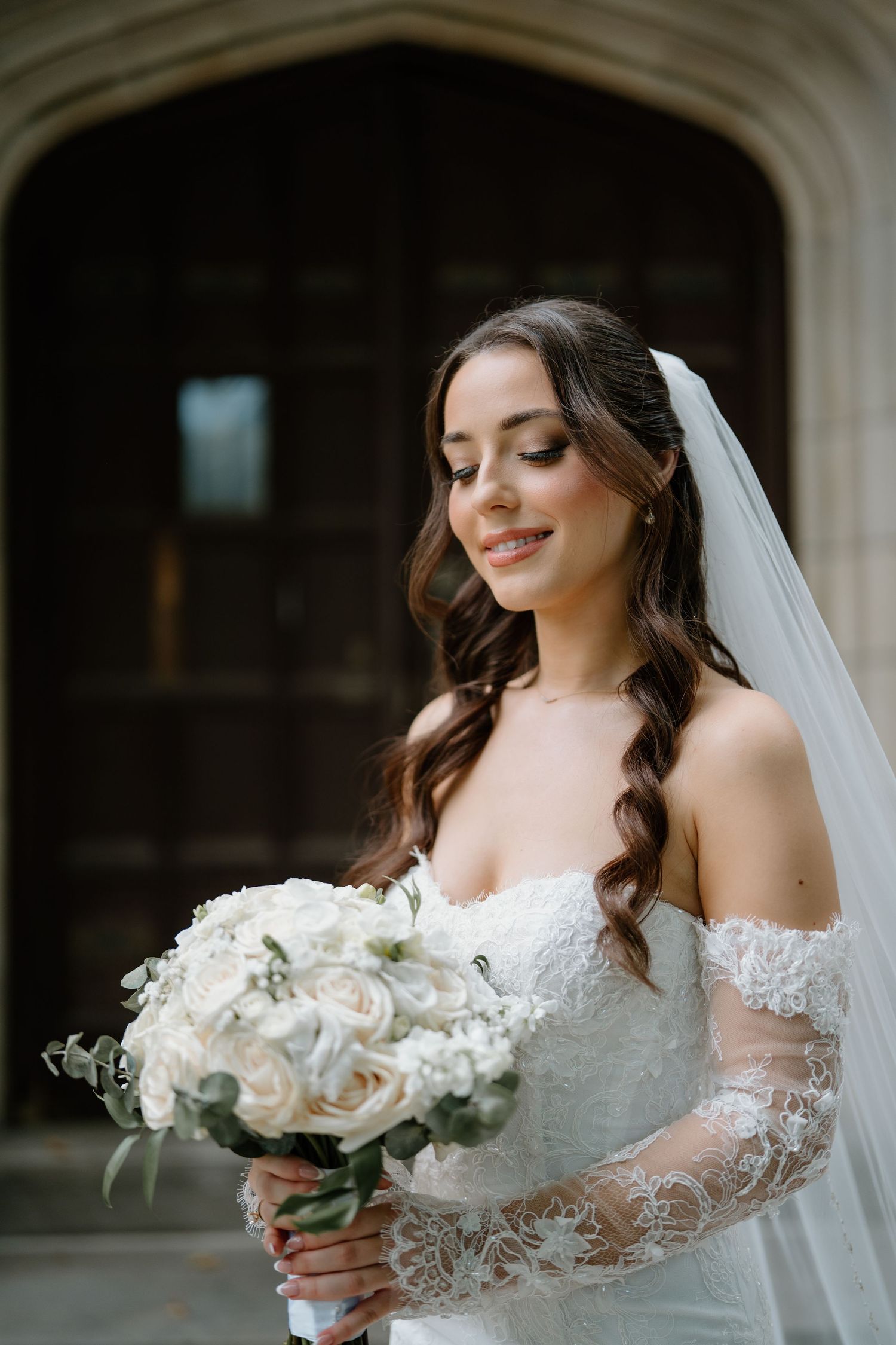 A bride in a lace wedding dress holds an elegant white rose bouquet against an architectural backdrop.