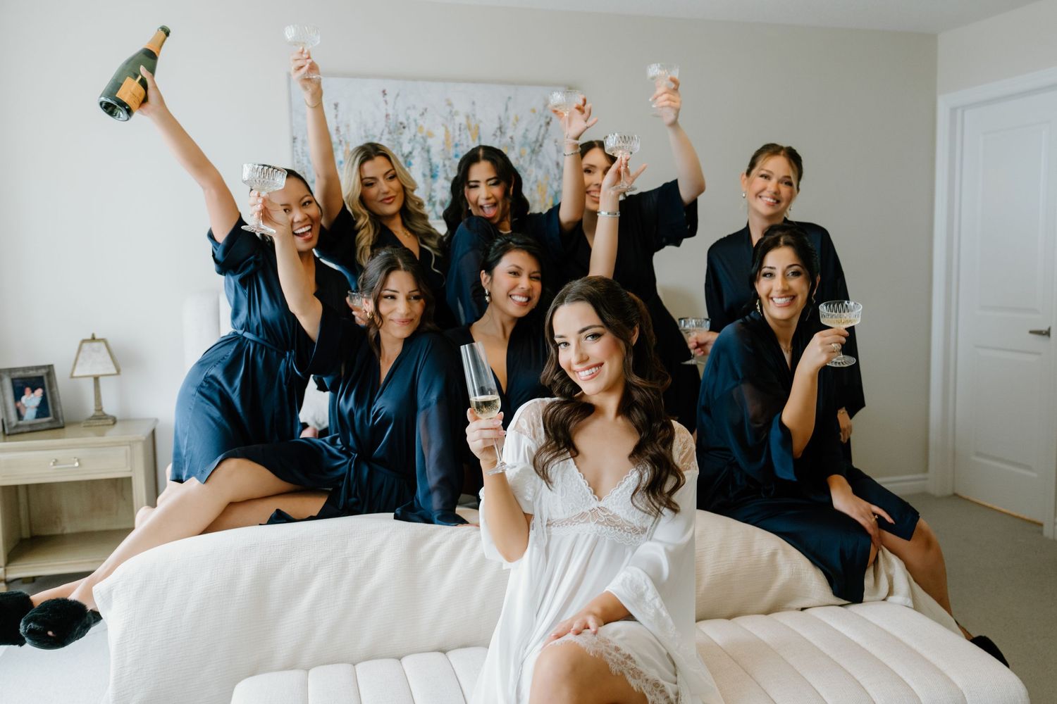 Bride and bridesmaids in navy blue dresses pose playfully together on a bed in a bright room.