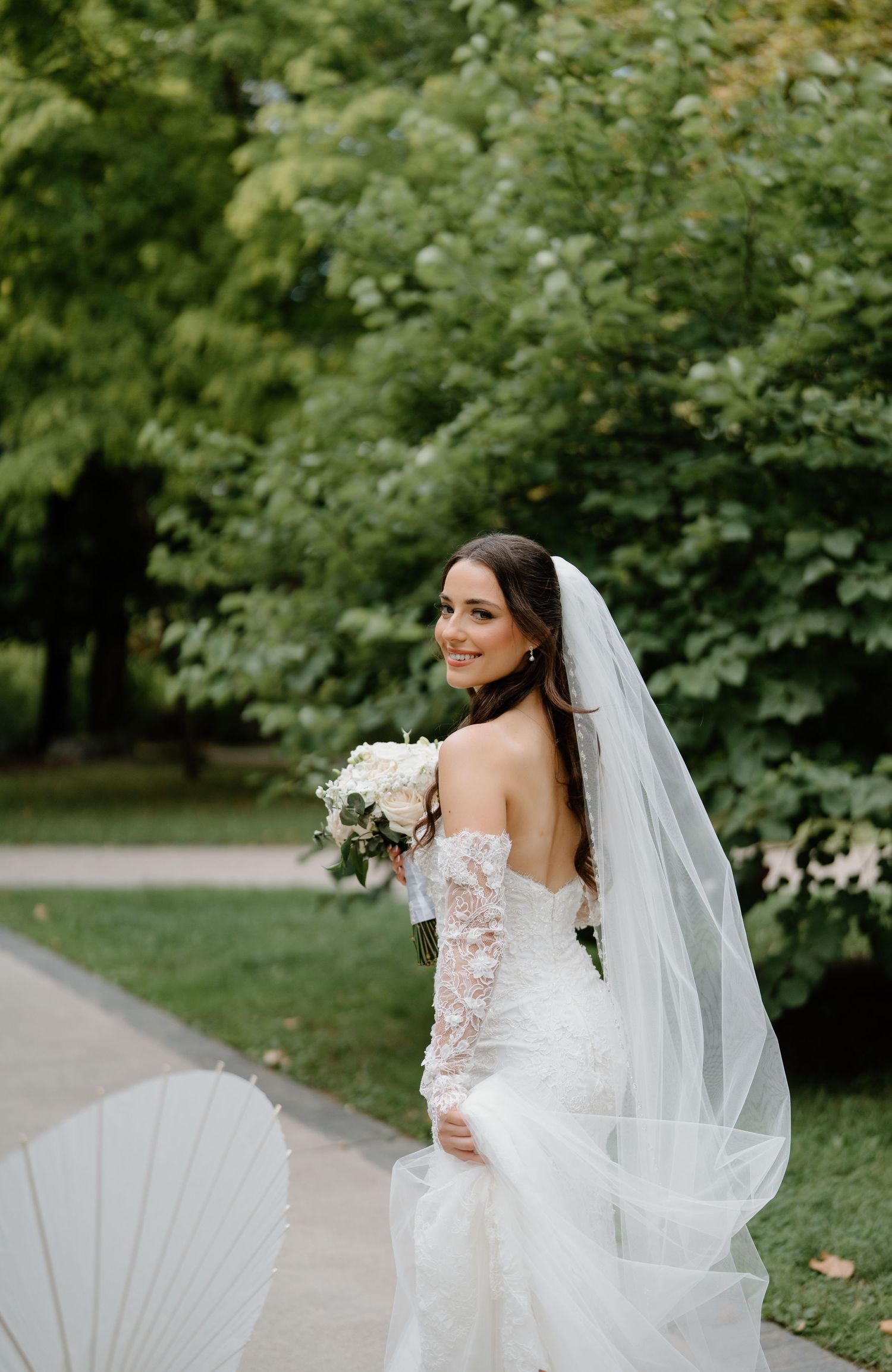A bride in a white lace gown with flowing veil poses elegantly in a lush garden setting.