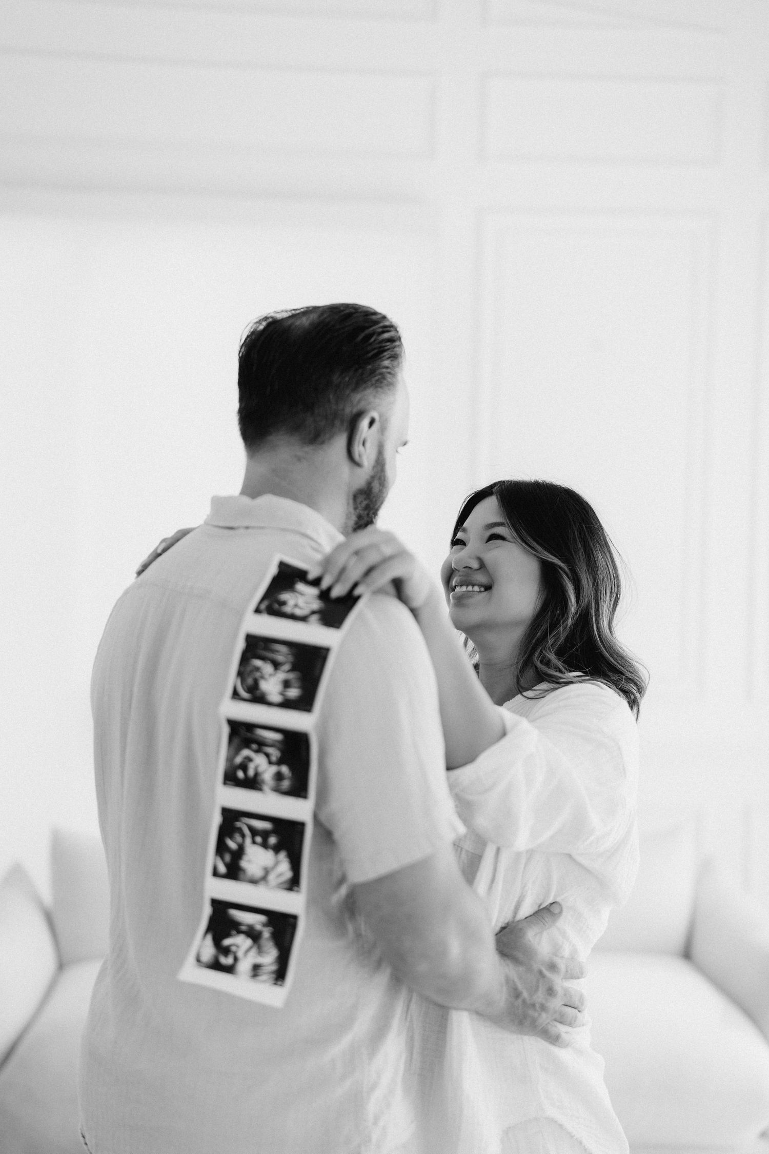 Couple in white clothing shares intimate moment with ultrasound photos displayed on their backs.