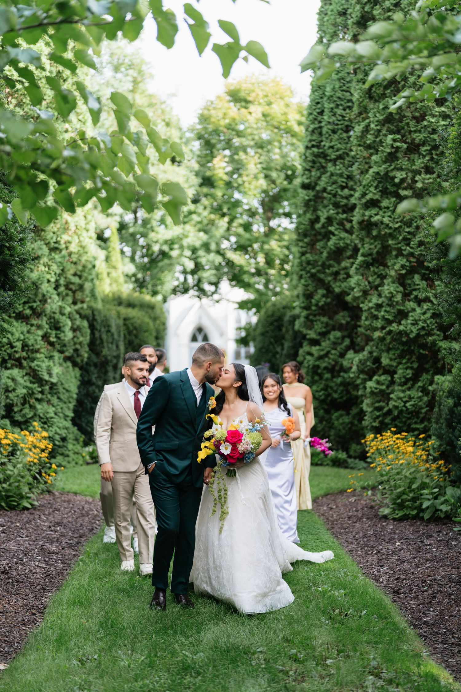 A wedding party walks along a manicured garden path with lush greenery and a white gazebo in the background.