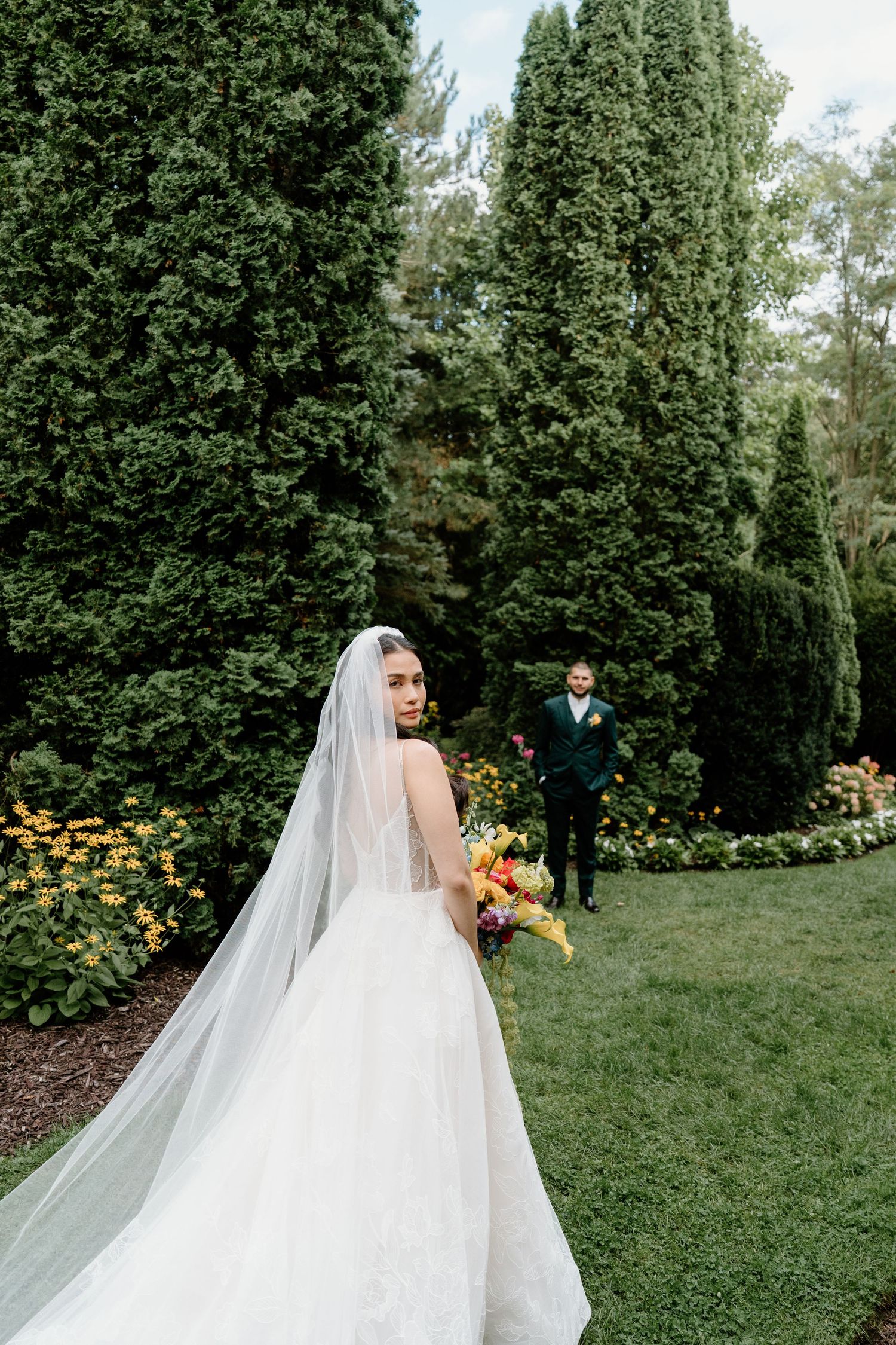 A couple shares an intimate moment in a garden setting with lush greenery and yellow flowers in the background.