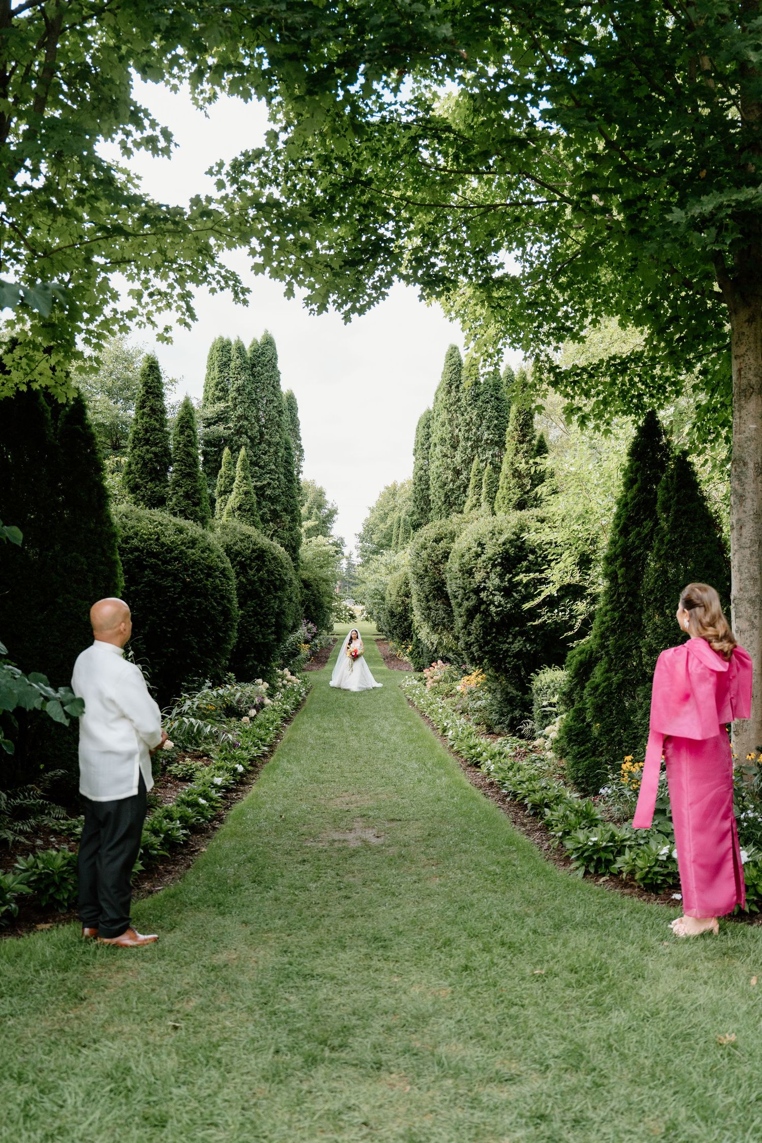 A manicured garden path lined with tall evergreen hedges leads to a distant ceremony setup.