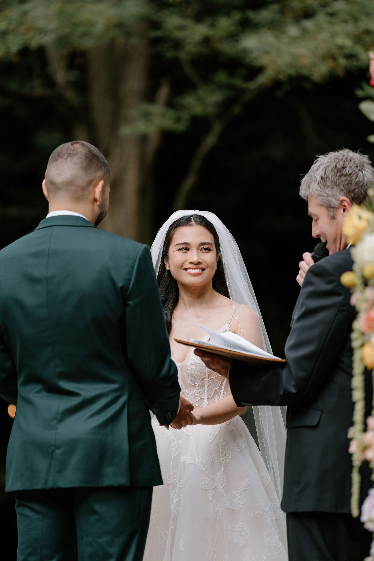 A wedding ceremony takes place outdoors with a couple and officiant standing beneath trees.