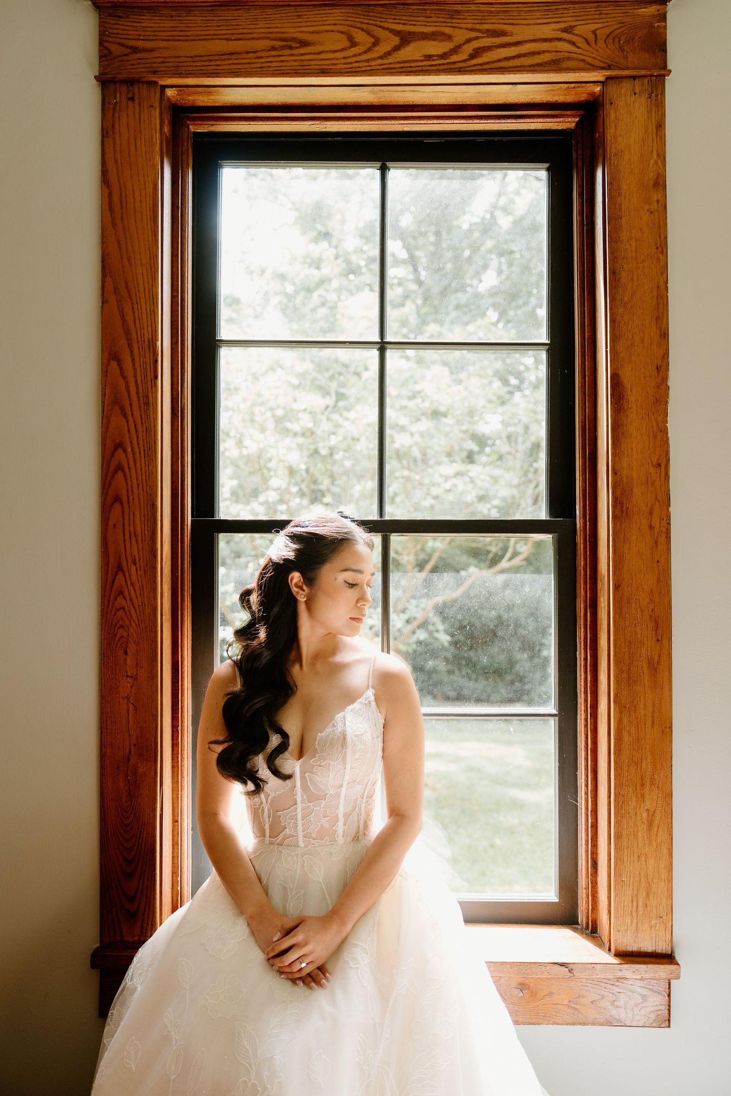 A bride in a white wedding gown sits elegantly by a window with wooden trim in natural lighting.