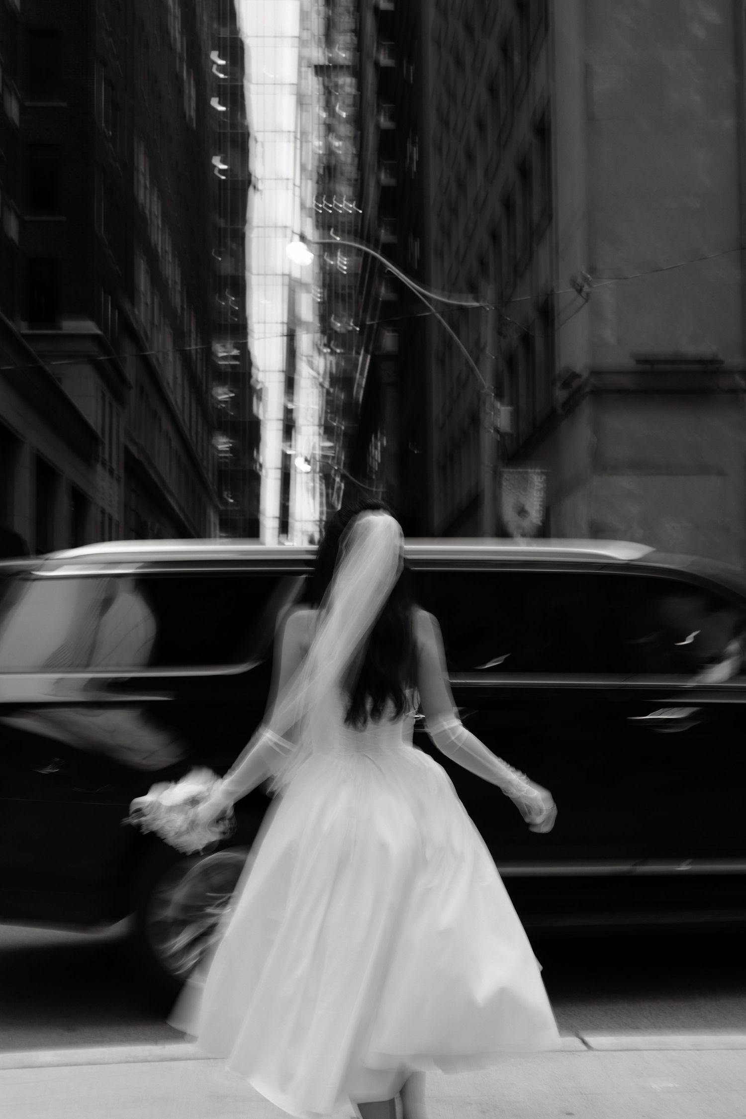 A blurred motion black and white photograph of a person in a white dress twirling on a city street with cars in background.